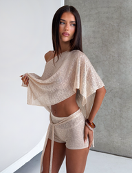 A woman with long dark hair models TIGERMIST’s FERN CROCHET SHORT in Champagne, styled with a sheer knit off-the-shoulder top. She stands against a textured gray wall, lifting part of the top and resting her hand on her hip.