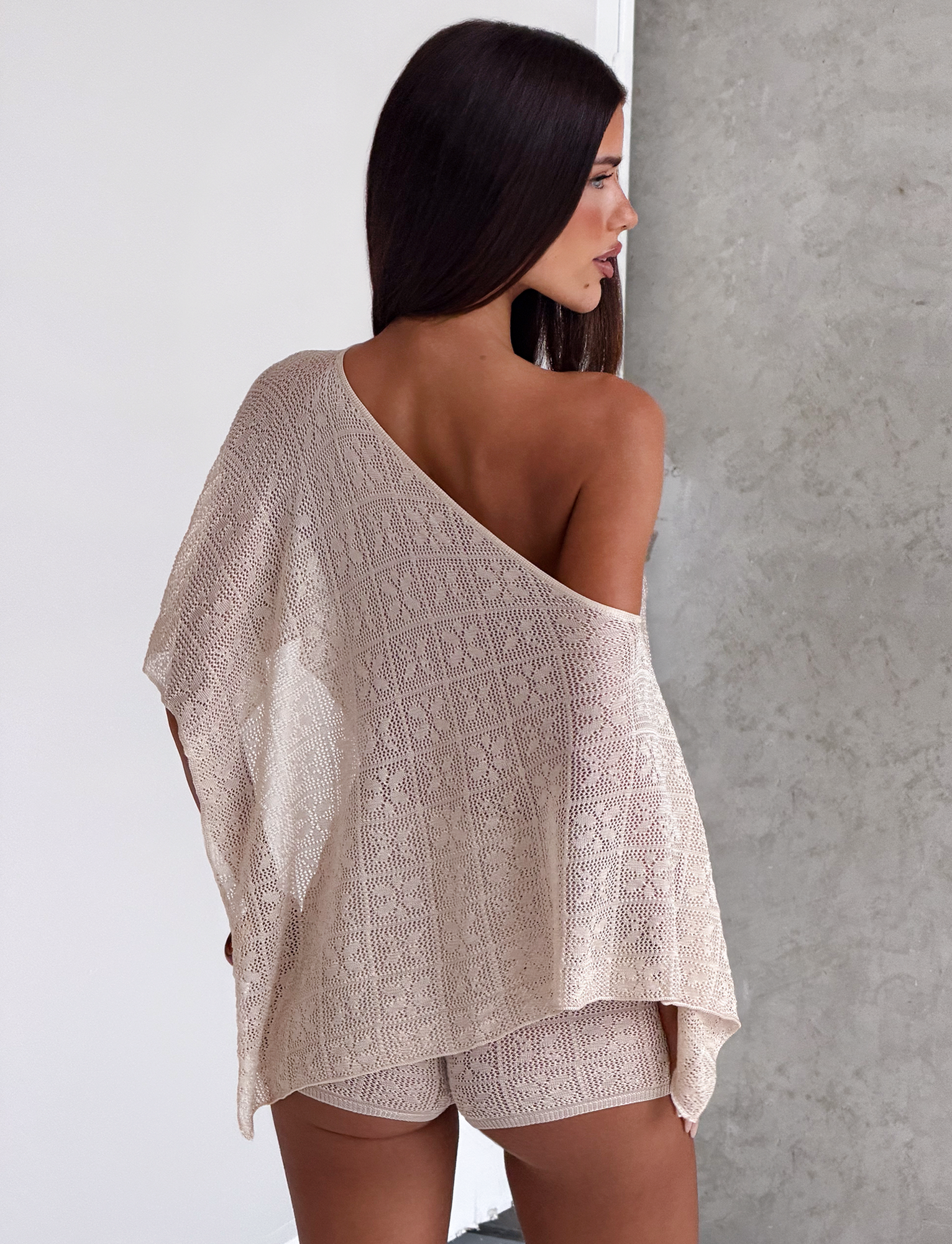 A woman with long dark hair stands sideways in a minimalist room, wearing TIGERMIST's FERN CROCHET SHORT in Champagne and a beige off-the-shoulder knit top.