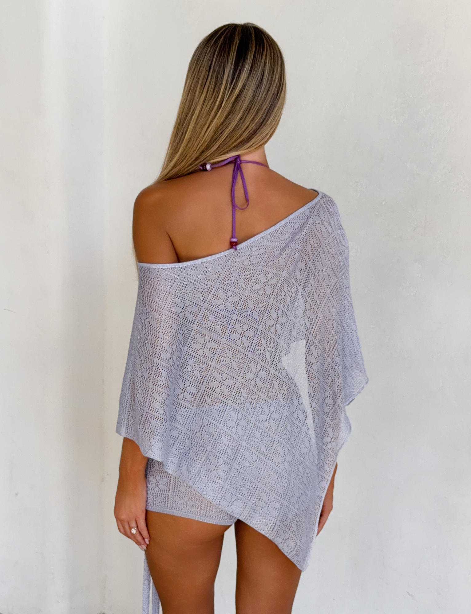 A woman with long, straight hair faces a white wall, wearing the TIGERMIST FERN CROCHET PONCHO in silver over a purple halter bikini top. The sheer cover-up drapes off one shoulder, showcasing her upper back.