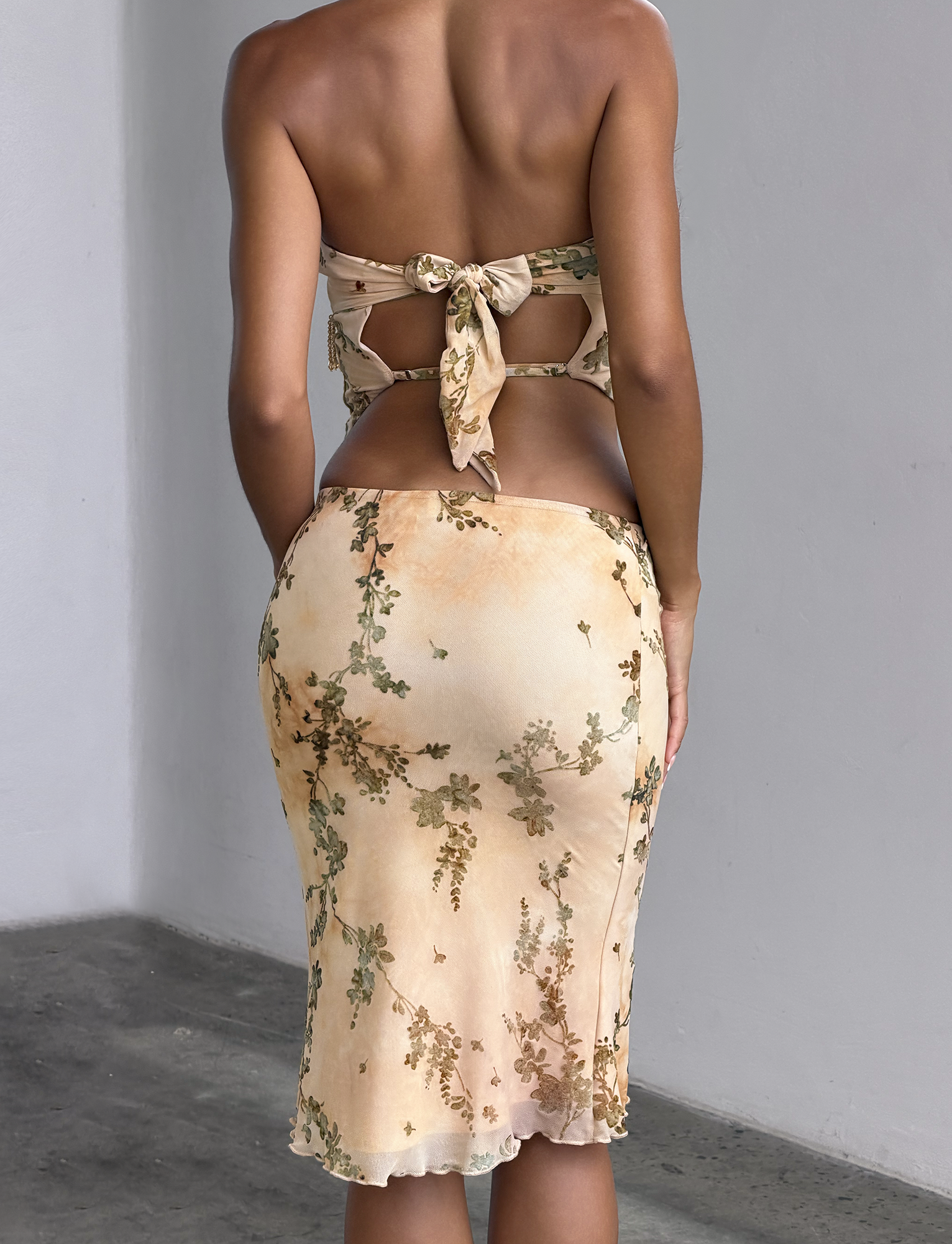 A woman stands with her back to the camera, wearing the TIGERMIST OONA BURN OUT FRINGE TOP in tan, featuring a floral pattern, tied straps, and beaded fringe trim. She is indoors by a white wall and gray concrete floor.
