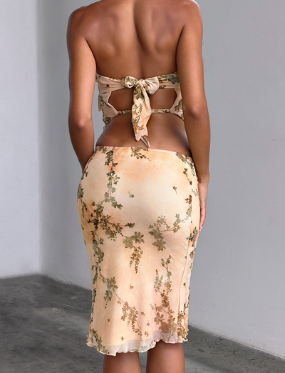 A person stands indoors with their back to the camera, wearing the TIGERMIST OONA BURN OUT FRINGE SKIRT in tan, featuring a light fringe texture. The setting is simple, with plain white walls and a concrete floor.