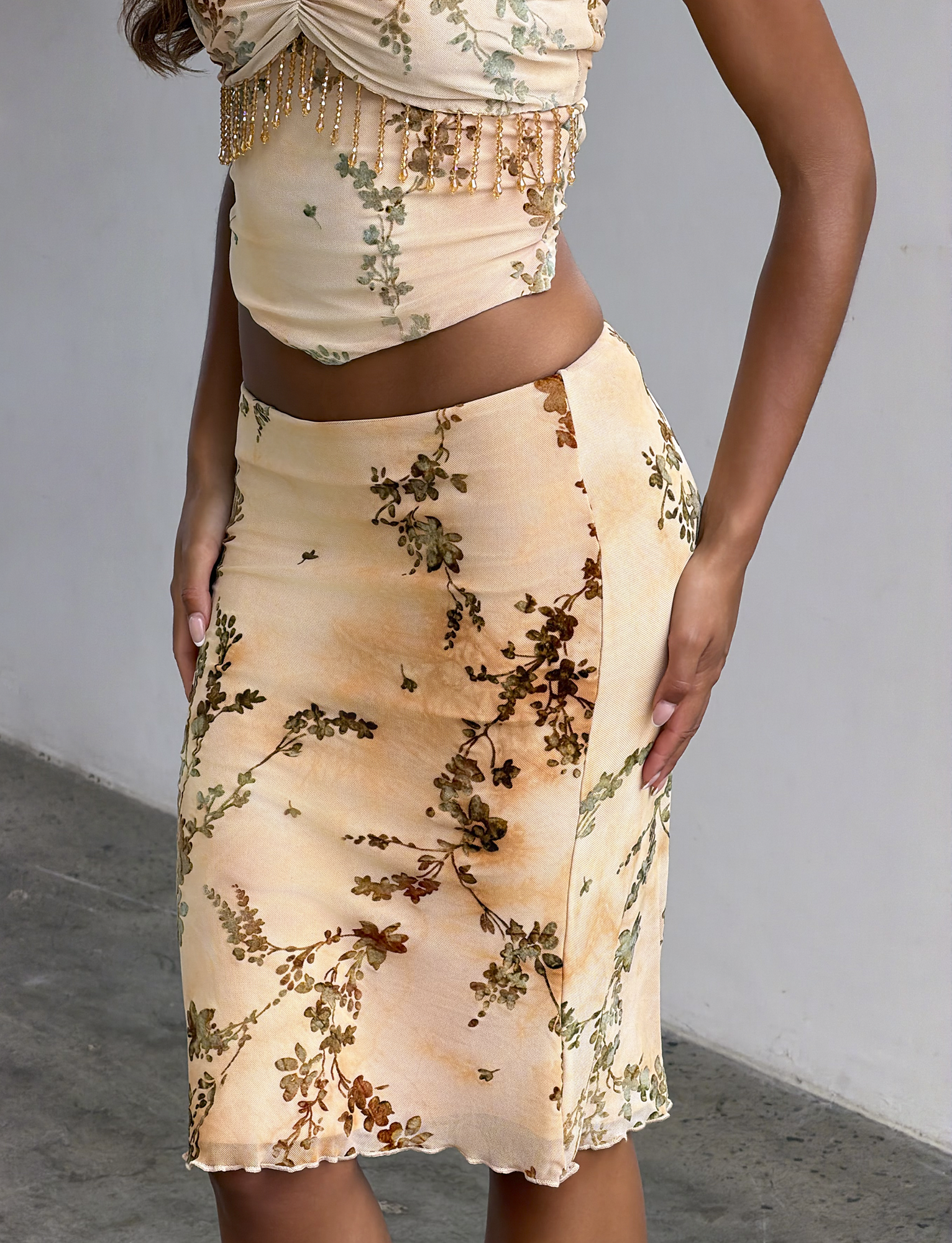 A person wearing the TIGERMIST OONA BURN OUT FRINGE SKIRT in tan with a fitted, beaded crop top stands against a plain background.