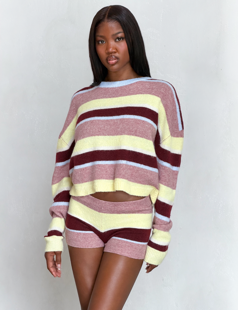 A woman with long, straight hair and a neutral expression wears the TIGERMIST KOA 2.0 STRIPED JUMPER in tan stripe, posing against a light gray background.