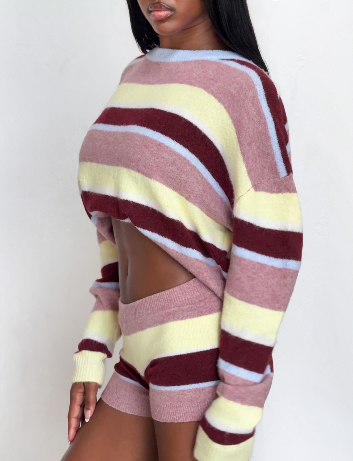 A woman wears TIGERMIST’s KOA 2.0 STRIPED SHORT - TAN STRIPE, a chic matching knit set with multicolour stripes in blush pink, burgundy, pale yellow, and light blue, pairing a cropped sweater with stripe knit mini shorts for a coordinated look.