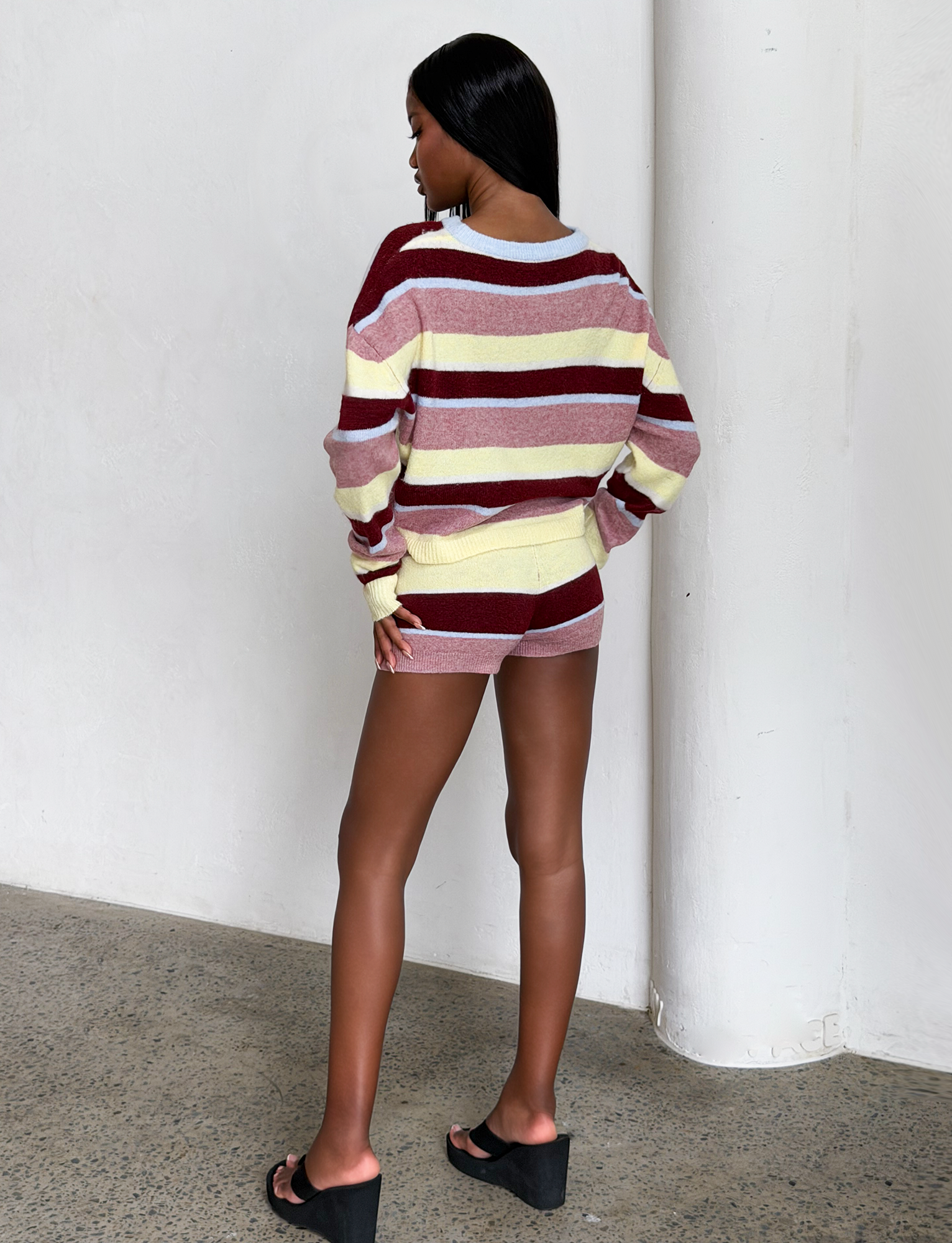 A woman with straight dark hair faces away from the camera, wearing the TIGERMIST KOA 2.0 STRIPED JUMPER in Tan Stripe with black platform sandals, standing against a plain white wall.