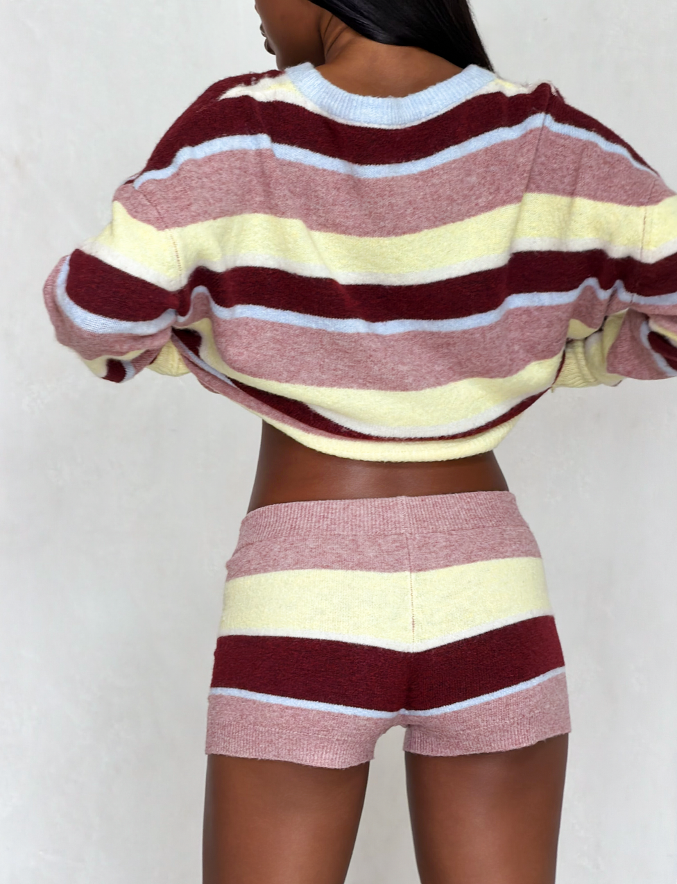 A woman stands with her back to the camera, wearing a cozy knit sweater and TIGERMIST KOA 2.0 STRIPED SHORT - TAN STRIPE featuring bold maroon, yellow, pink, and white stripes that stand out against a plain light-colored wall.