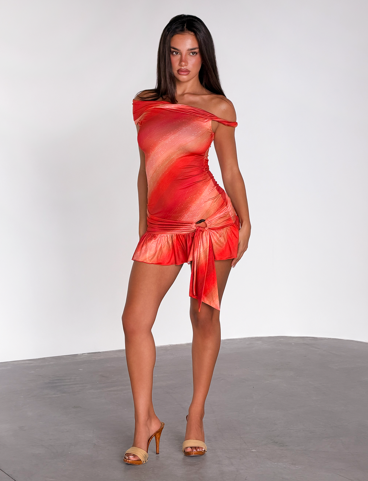A woman with long dark hair and a neutral expression stands on a gray floor, wearing the TIGERMIST HAELO MINI DRESS in Sunrise Glitter Stripe and beige high-heeled sandals against a plain white background.