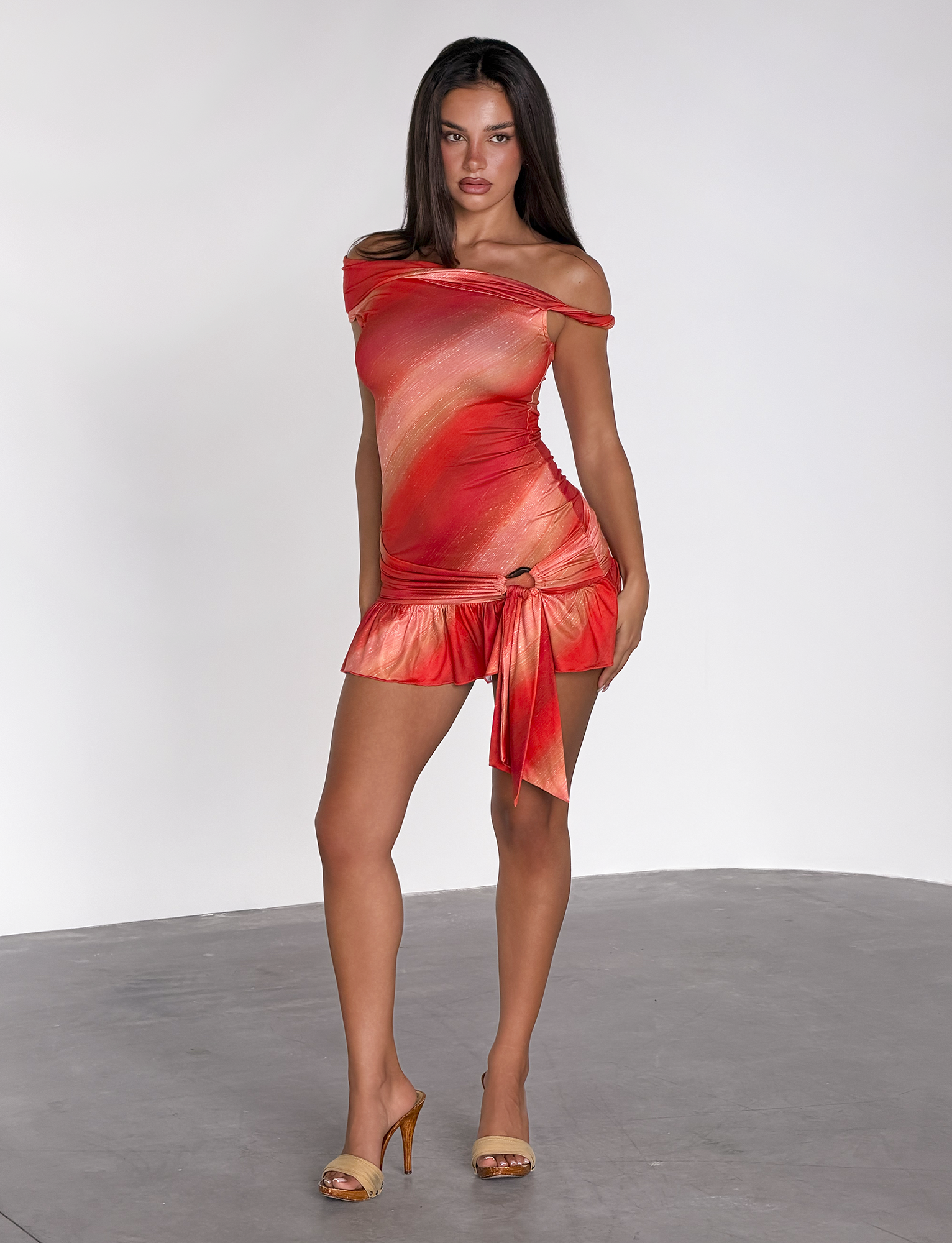 A woman with long dark hair and a neutral expression stands on a gray floor, wearing the TIGERMIST HAELO MINI DRESS in Sunrise Glitter Stripe and beige high-heeled sandals against a plain white background.