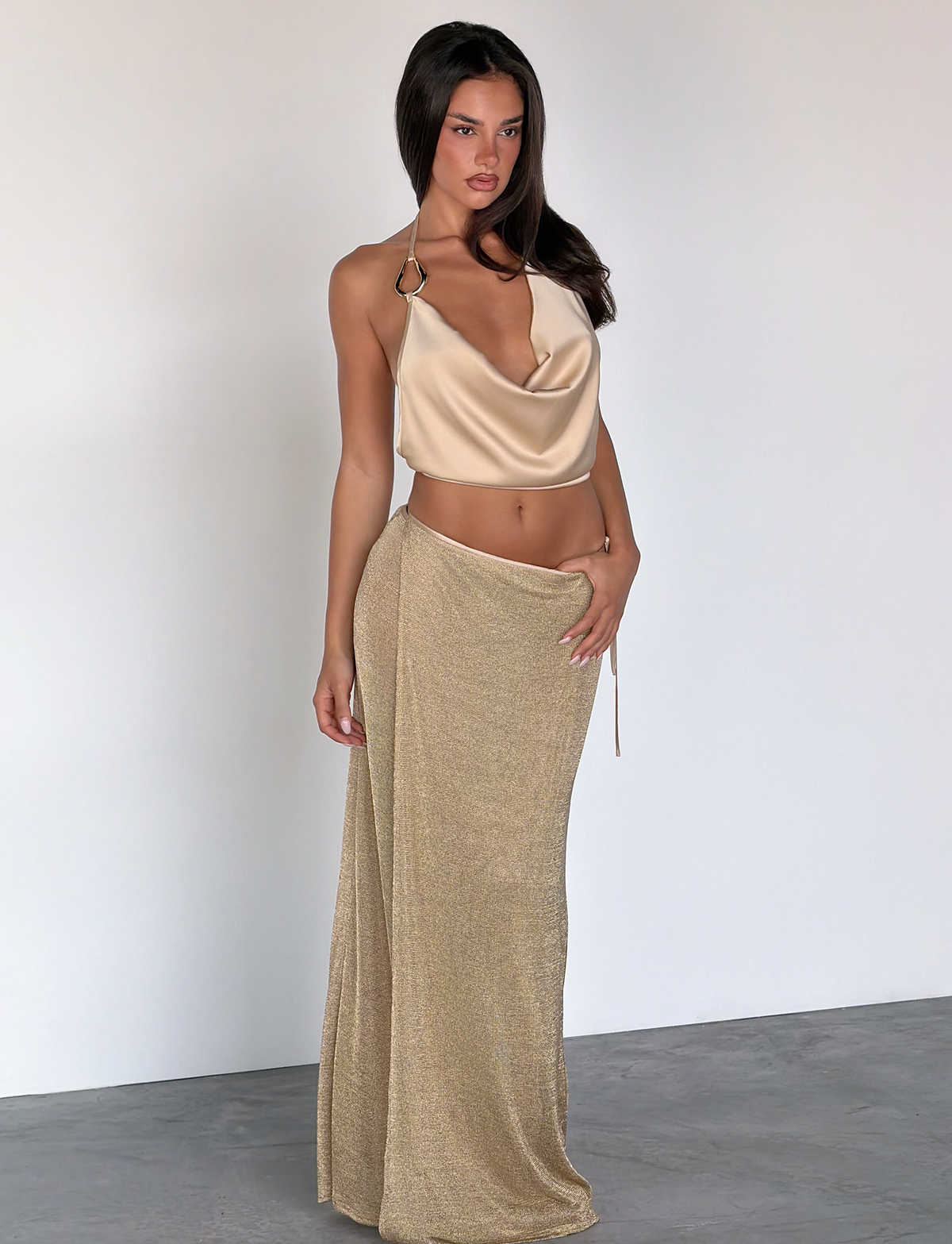 A woman with long brown hair and a neutral expression stands against a white wall, wearing the TIGERMIST CYRENE TIE MAXI SKIRT - GOLD paired with a silky gold halter top. One hand rests on her hip.