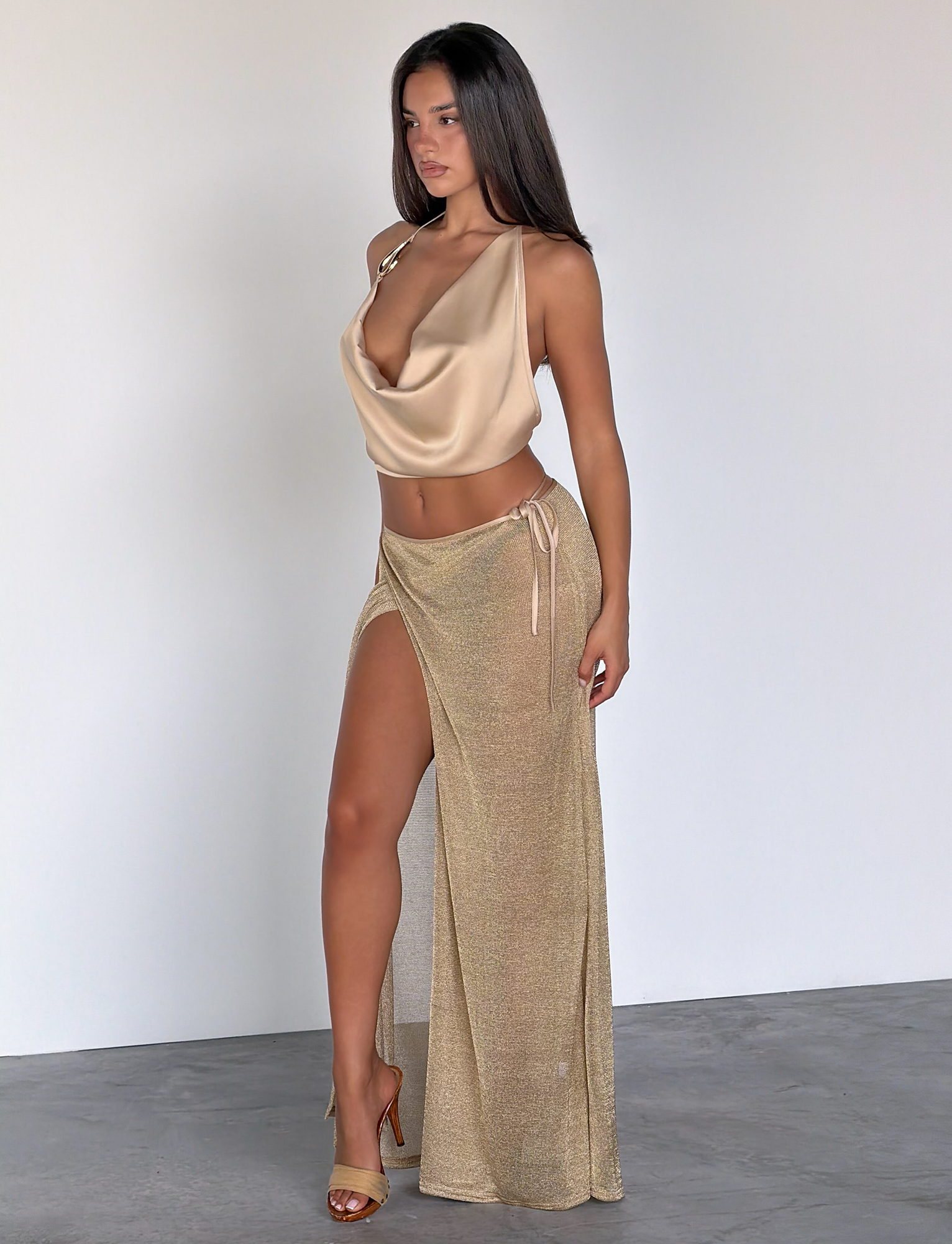 A woman with long dark hair wears the TIGERMIST CYRENE TIE MAXI SKIRT in gold—a high-slit, semi-sheer metallic knit skirt—paired with a champagne cowl-neck top and heels, standing indoors against a white wall, looking slightly to the side.