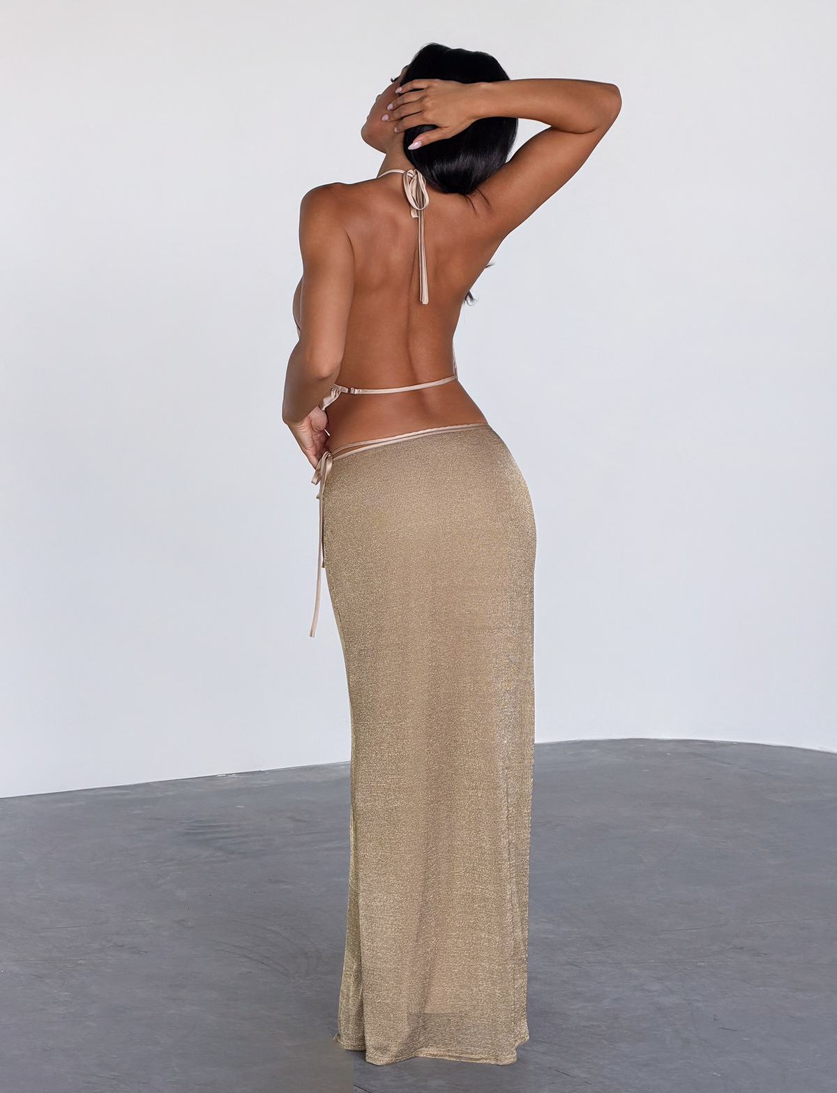 A woman with long dark hair stands barefoot on a gray floor, facing away. She poses in the TIGERMIST CYRENE SATIN COWL TOP in gold, against a plain light background, with one hand on her head and the other on her hip.