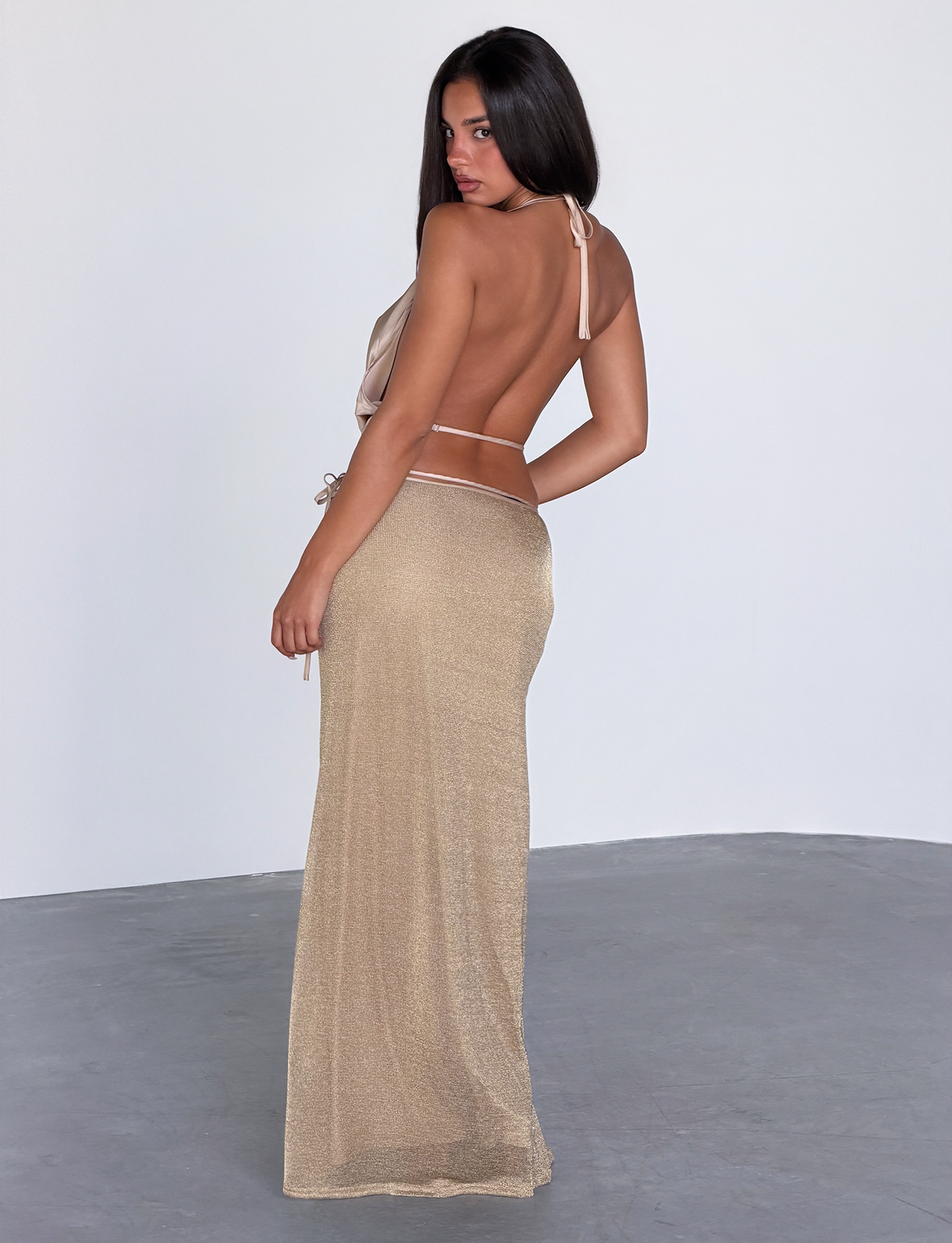 A woman with long dark hair stands in a minimalist room wearing the TIGERMIST CYRENE TIE MAXI SKIRT in gold, looking over her shoulder at the camera.