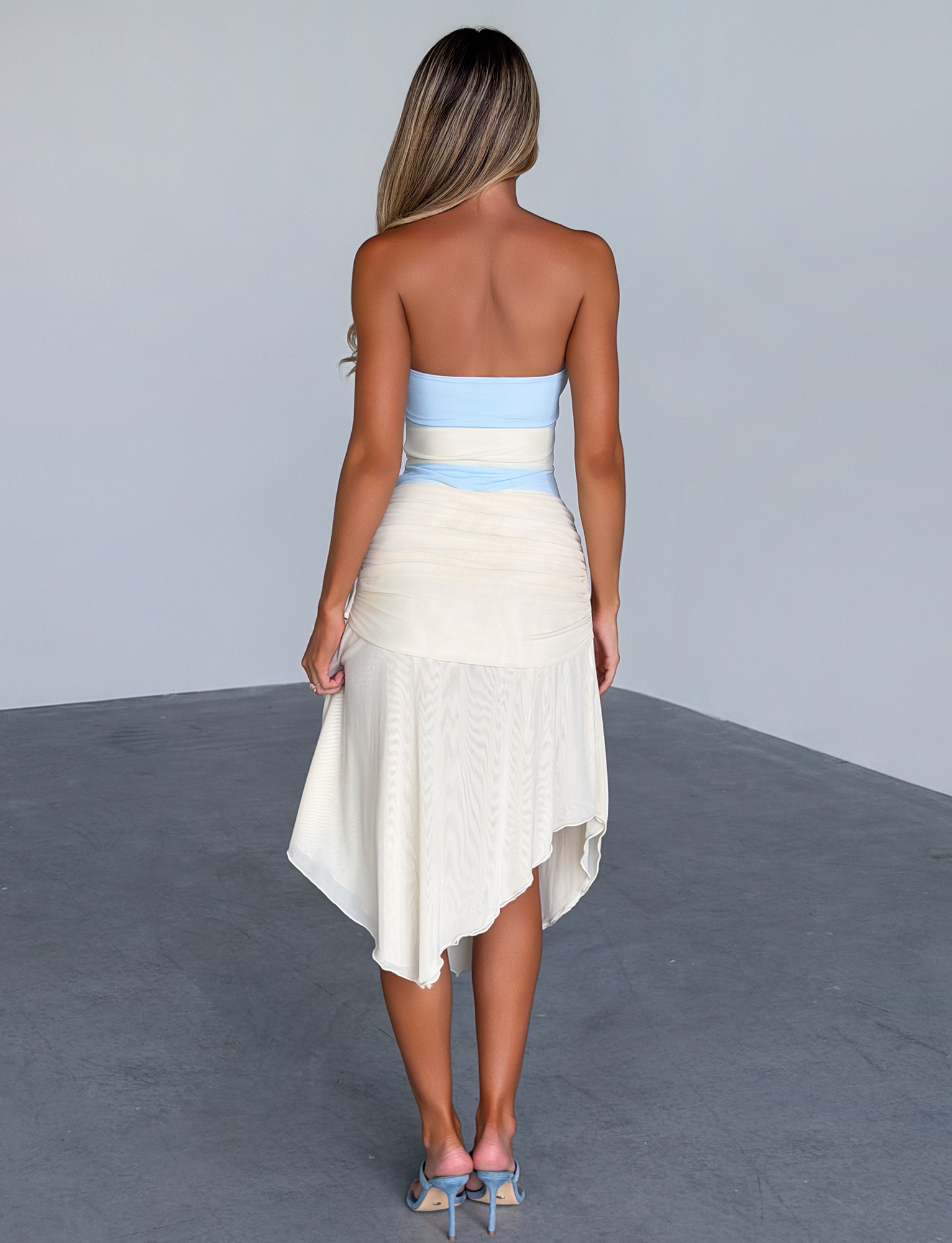 A woman with long blonde hair stands indoors facing away, wearing the TIGERMIST TWYLA MIDI DRESS in cream and blue, on a gray concrete floor with a plain pale wall background.