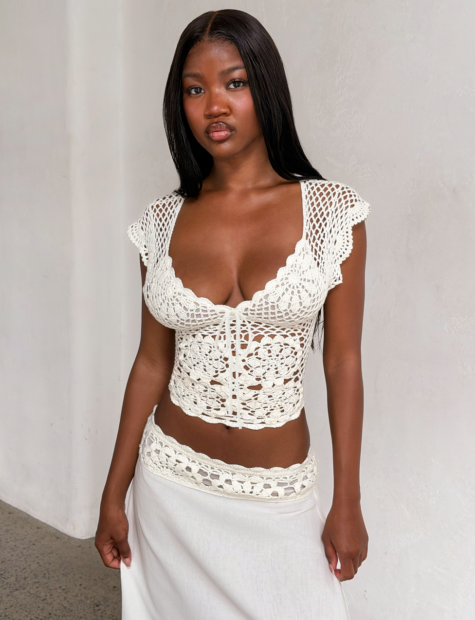 A woman with long, straight hair wears the TIGERMIST SASHA CROCHET TOP in ivory, featuring scalloped cap sleeves, and stands against a plain white wall while looking at the camera.