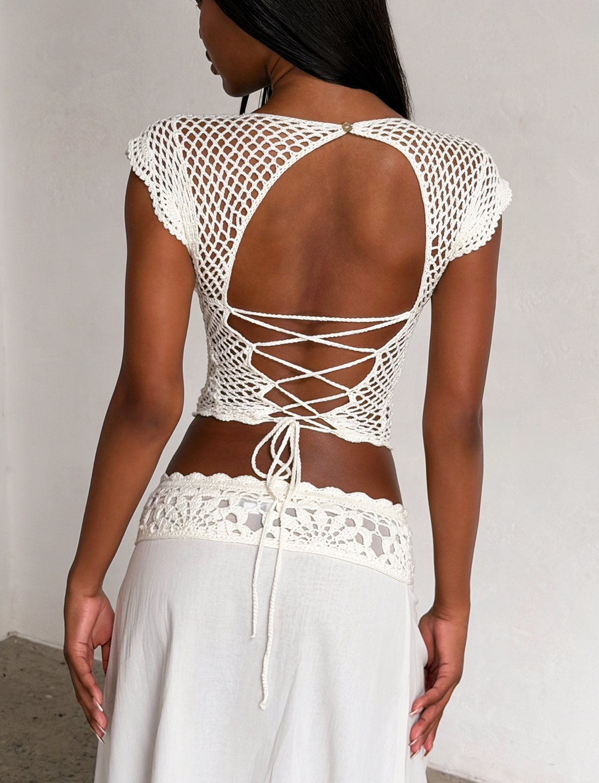 A woman stands facing away, wearing the TIGERMIST SASHA CROCHET TOP in ivory with scalloped cap sleeves and crisscross back, paired with a white crochet-trim skirt. The summery outfit is set against a plain background.