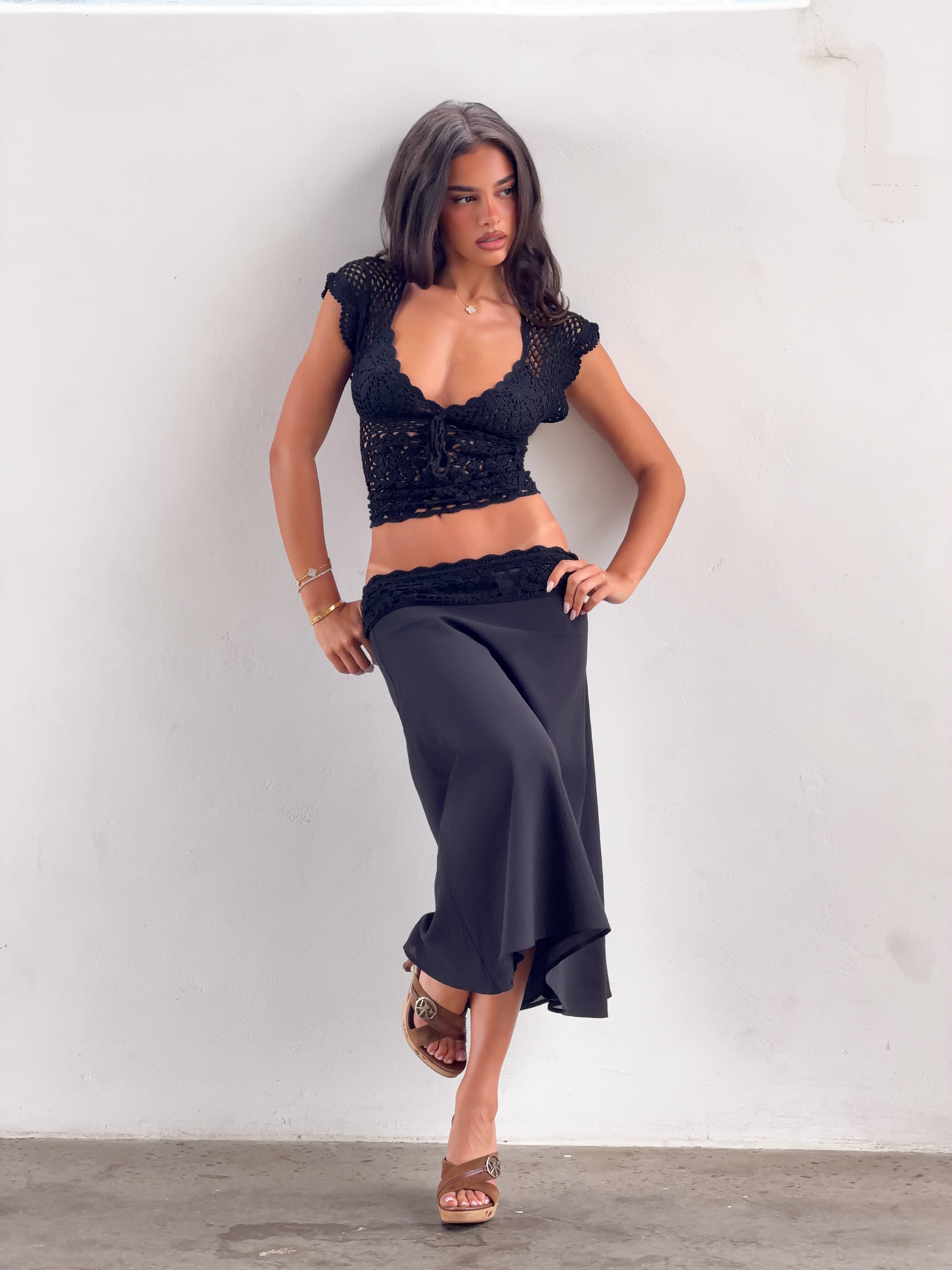 A woman with long dark hair poses against a white wall in the TIGERMIST SASHA CROCHET TOP - BLACK, paired with a long black skirt and brown heeled sandals. She stands relaxed, one leg crossed over the other—effortless resort style.