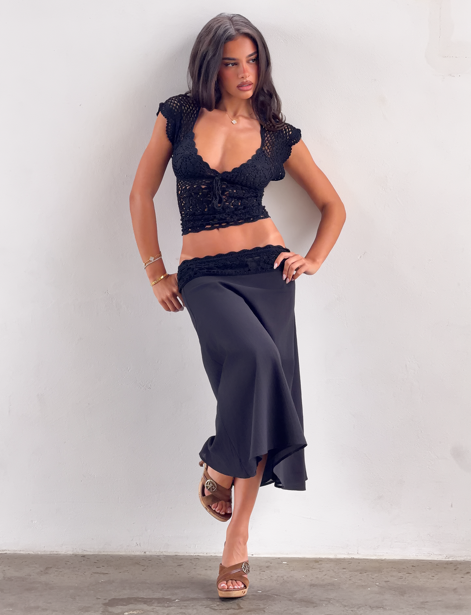 A woman poses against a white wall in the SASHA CROCHET MIDI SKIRT - BLACK by TIGERMIST, styled with a black crochet crop top and wedge sandals, with one hand on her hip.