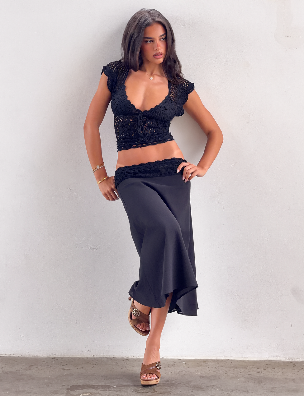 A woman poses against a white wall in the SASHA CROCHET MIDI SKIRT - BLACK by TIGERMIST, styled with a black crochet crop top and wedge sandals, with one hand on her hip.