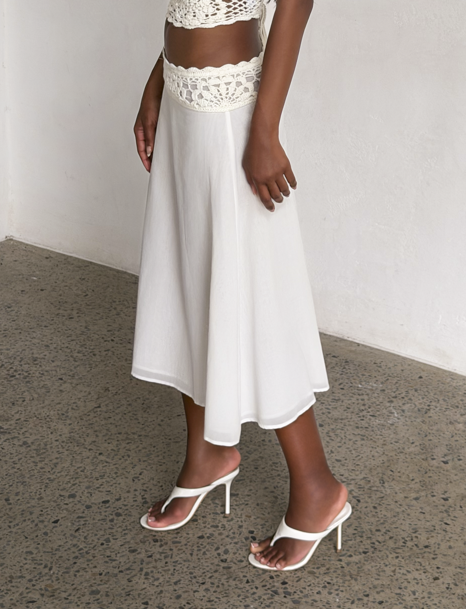 A person wearing the TIGERMIST SASHA MIDI SKIRT in ivory with a crochet waistband, a matching crochet top, and white heeled sandals stands on a speckled concrete floor against a plain white wall.