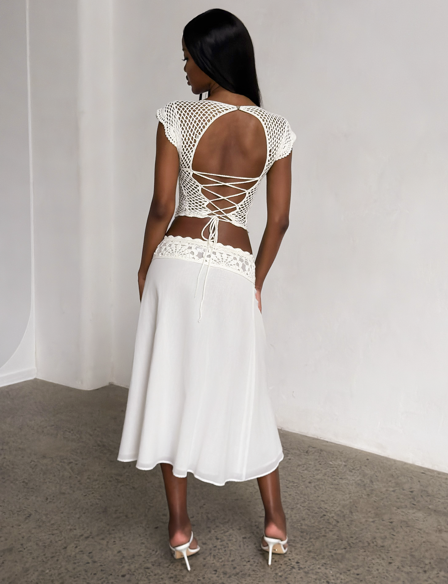 A woman stands indoors, facing away from the camera, wearing TIGERMIST’s SASHA MIDI SKIRT in ivory and a crochet-style open-back top with lace-up details, paired with high-heeled sandals on a polished concrete floor.