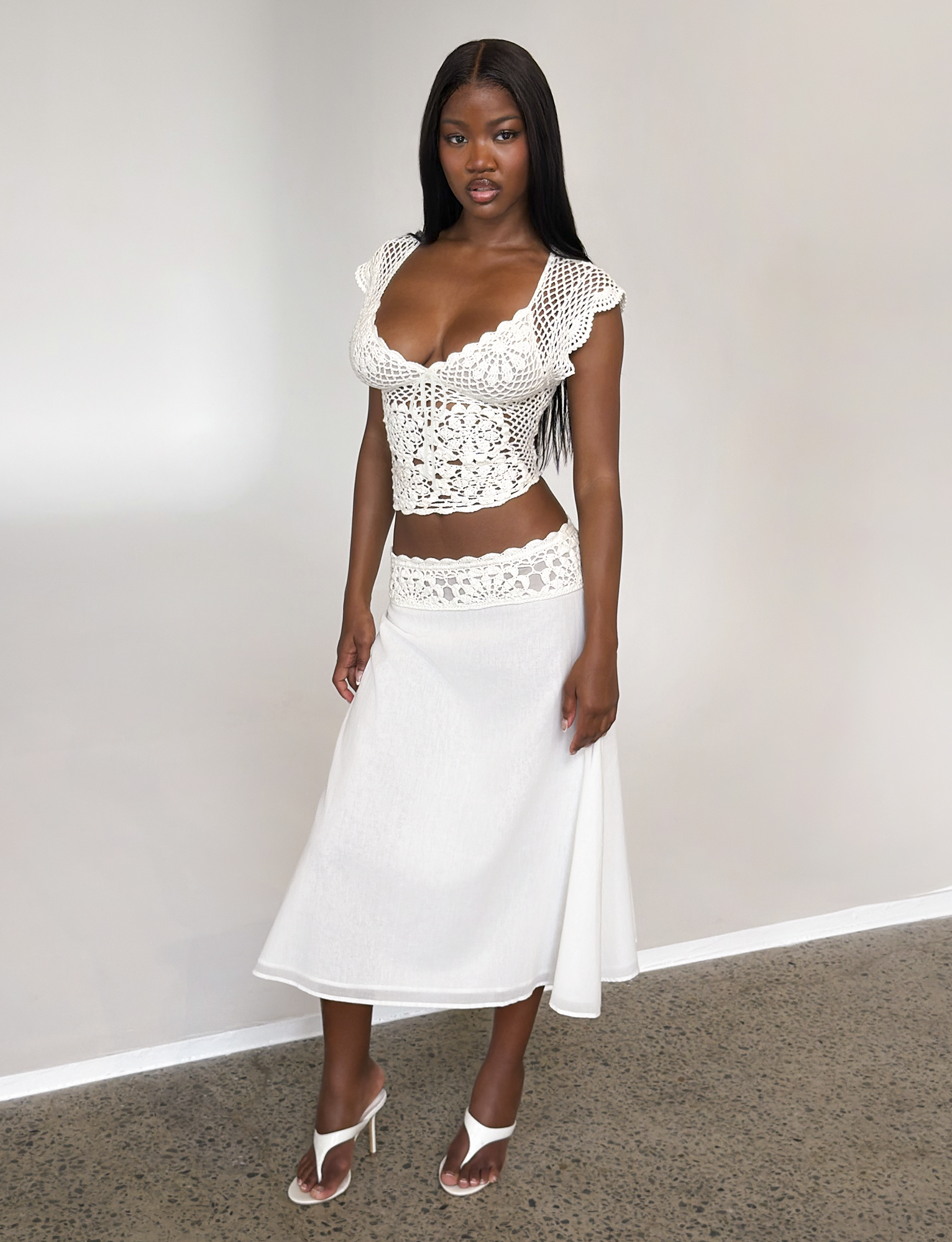 A woman stands indoors wearing the TIGERMIST SASHA MIDI SKIRT in ivory, paired with a white crocheted short-sleeve top featuring a deep neckline. She has long, straight hair and completes the look with white heeled sandals.