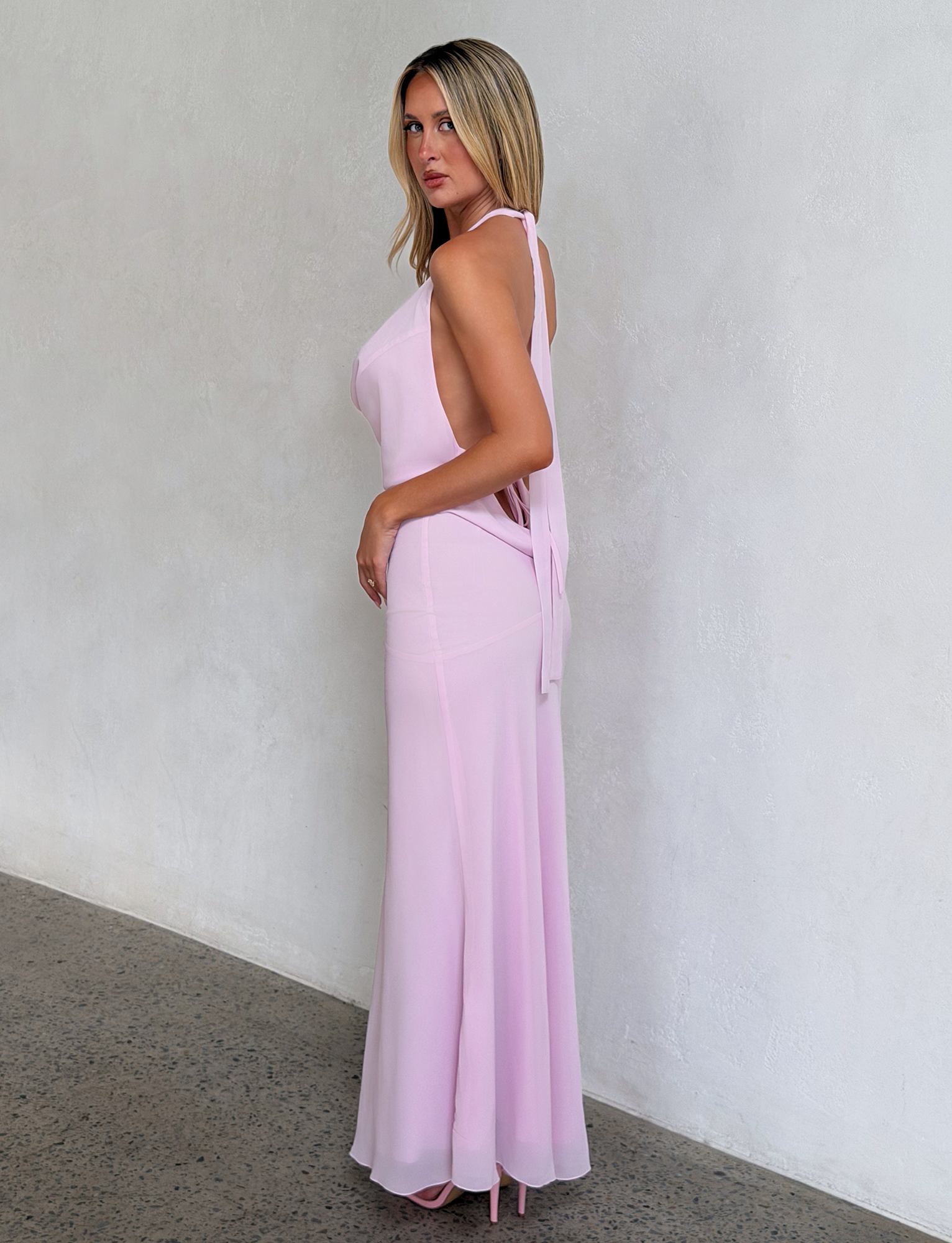 A woman with straight blonde hair stands sideways against a light textured wall, wearing the TIGERMIST ZYLA MAXI DRESS in baby pink and matching heels. She looks over her shoulder with a serious expression.