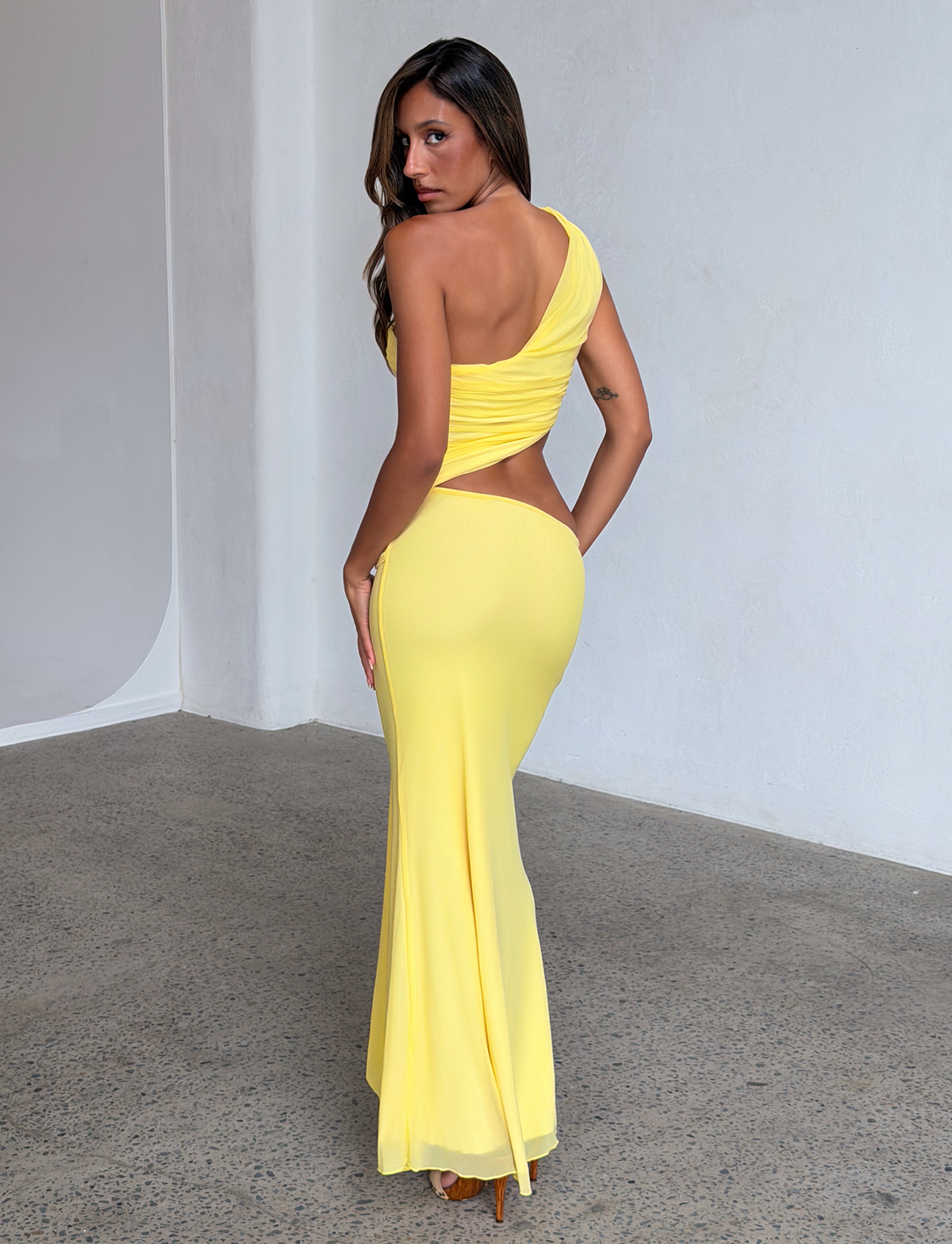 A woman poses indoors in the TIGERMIST COBI 2.0 MAXI DRESS in butter yellow, featuring a one-shoulder neckline and ruched bodice, standing on a speckled floor against a plain white wall.