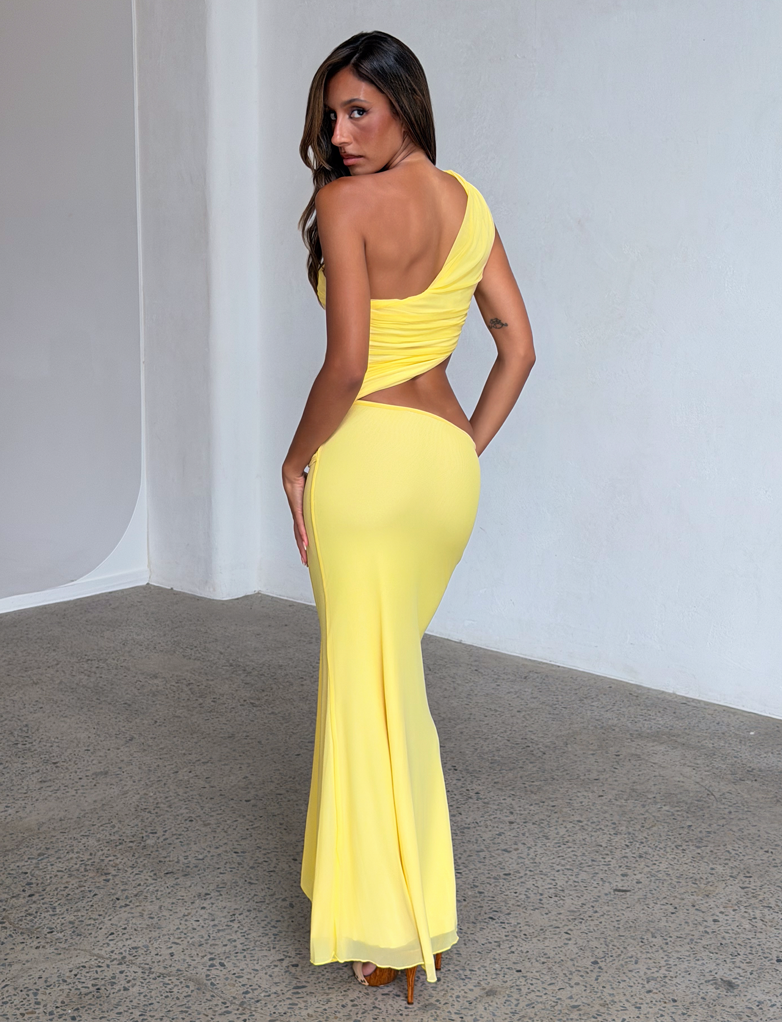 A woman poses indoors in the TIGERMIST COBI 2.0 MAXI DRESS in butter yellow, featuring a one-shoulder neckline and ruched bodice, standing on a speckled floor against a plain white wall.