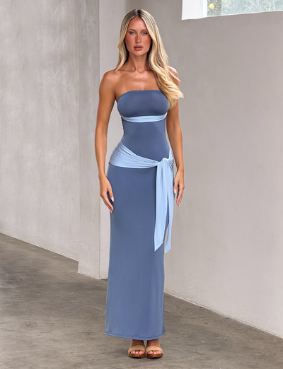 A woman poses indoors wearing the TIGERMIST TOVE MAXI DRESS - BLUE, a strapless, dark blue maxi dress with a lighter blue sash at the waist. She has long blonde hair and stands on a concrete floor against a neutral wall.