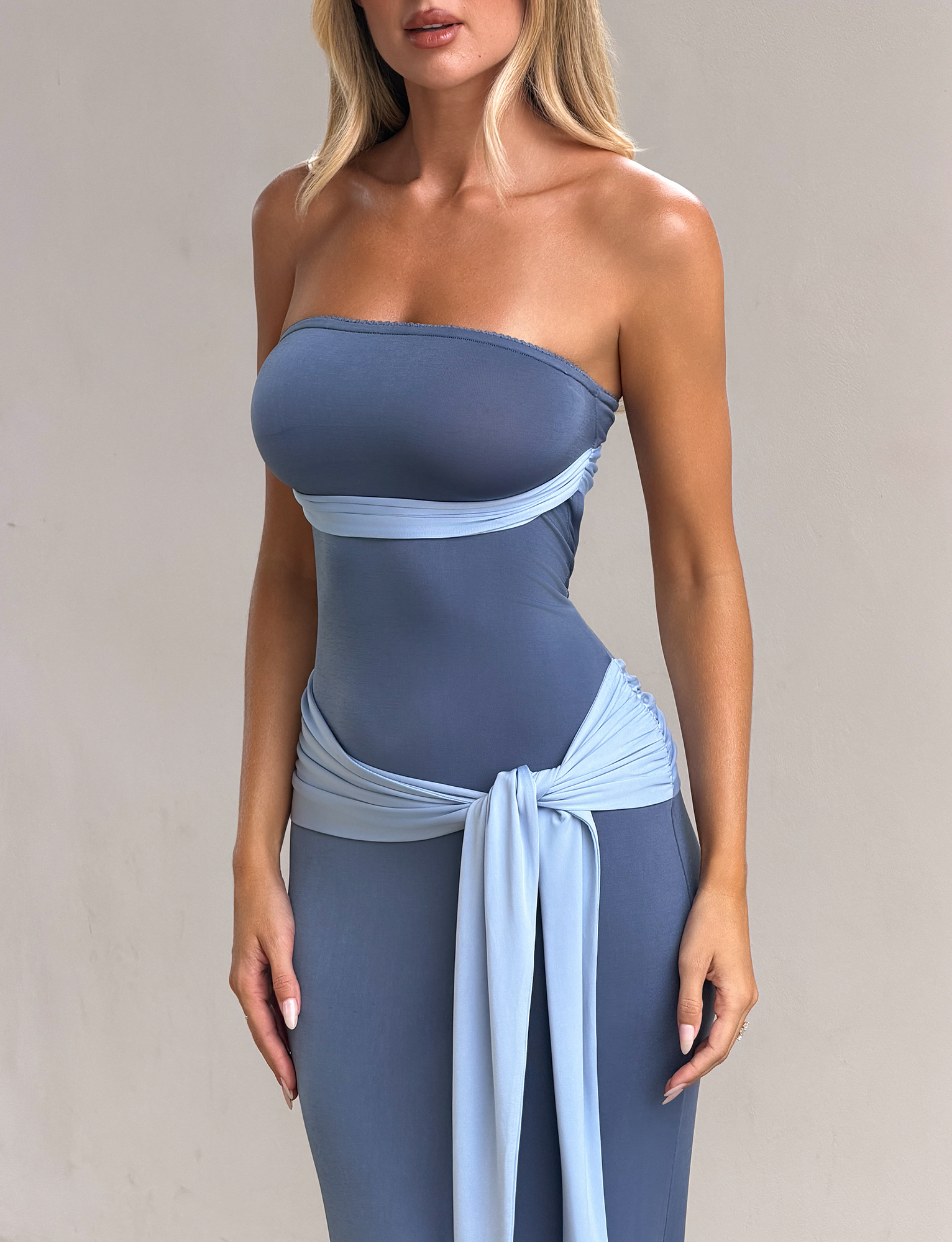 A woman models the TIGERMIST TOVE MAXI DRESS - BLUE, a strapless, fitted maxi dress with a light blue sash at the waist, standing against a plain light background. Her face is partially out of frame.