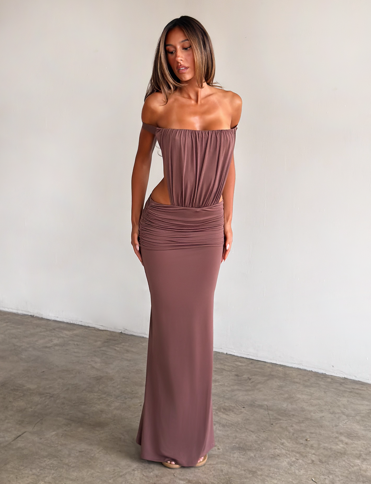 A woman with straight, shoulder-length hair stands indoors against a plain wall wearing the TIGERMIST IOLLA STRAPLESS MAXI DRESS in mocha, featuring a ruched bodice, cutout sides, and a fitted Grecian-style skirt.