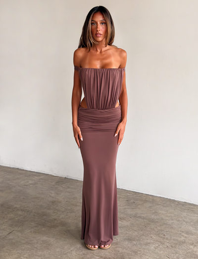 A woman stands against a plain light wall, wearing the TIGERMIST IOLLA STRAPLESS MAXI DRESS in mocha with an off-the-shoulder, ruched bodice and floor-length skirt. She has straight shoulder-length hair and neutral heels.
