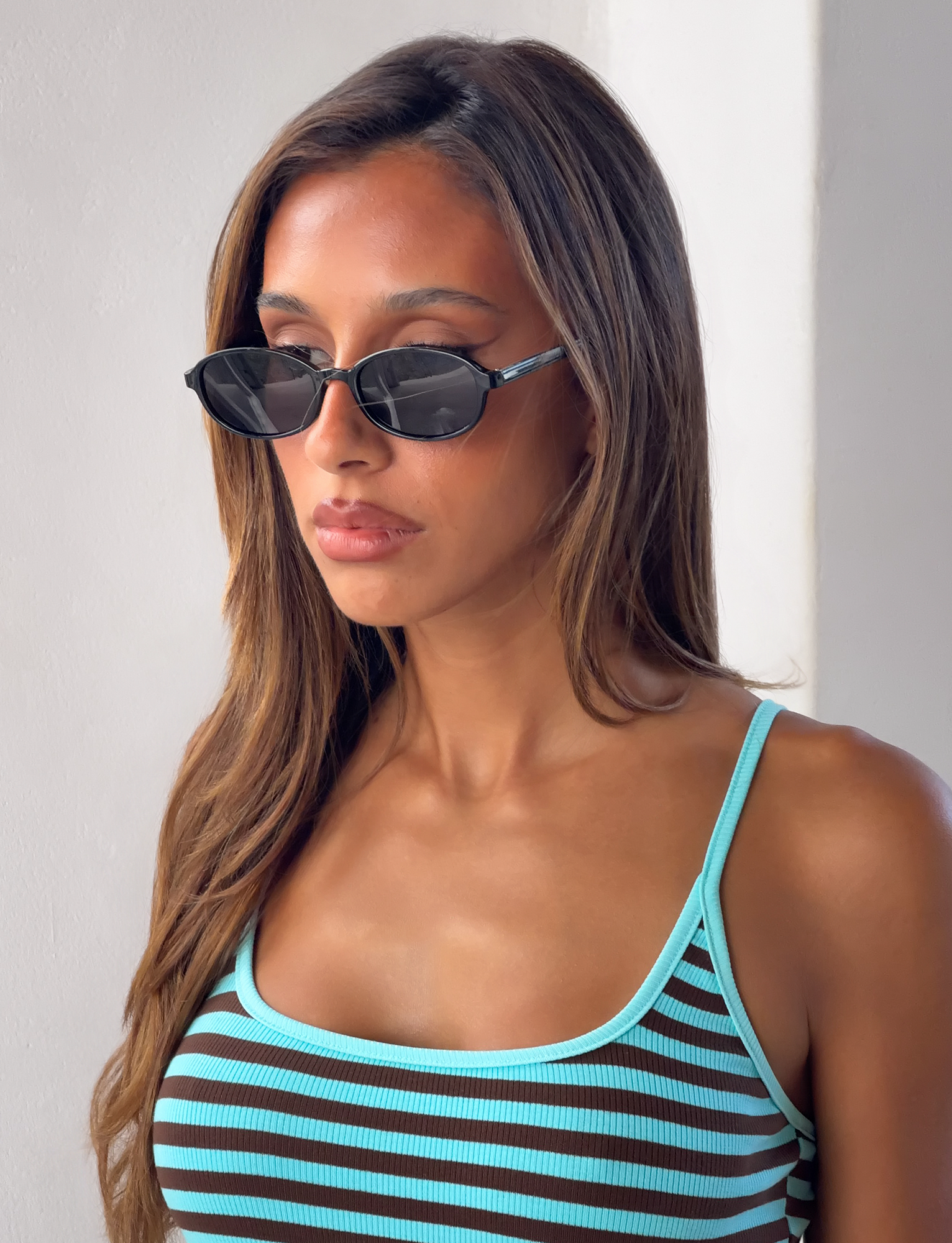 A woman with long brown hair wears Tiger Mist's CL - ESTA SUNGLASSES and a blue-striped tank top, standing in front of a white wall with a neutral expression, looking slightly to the side.