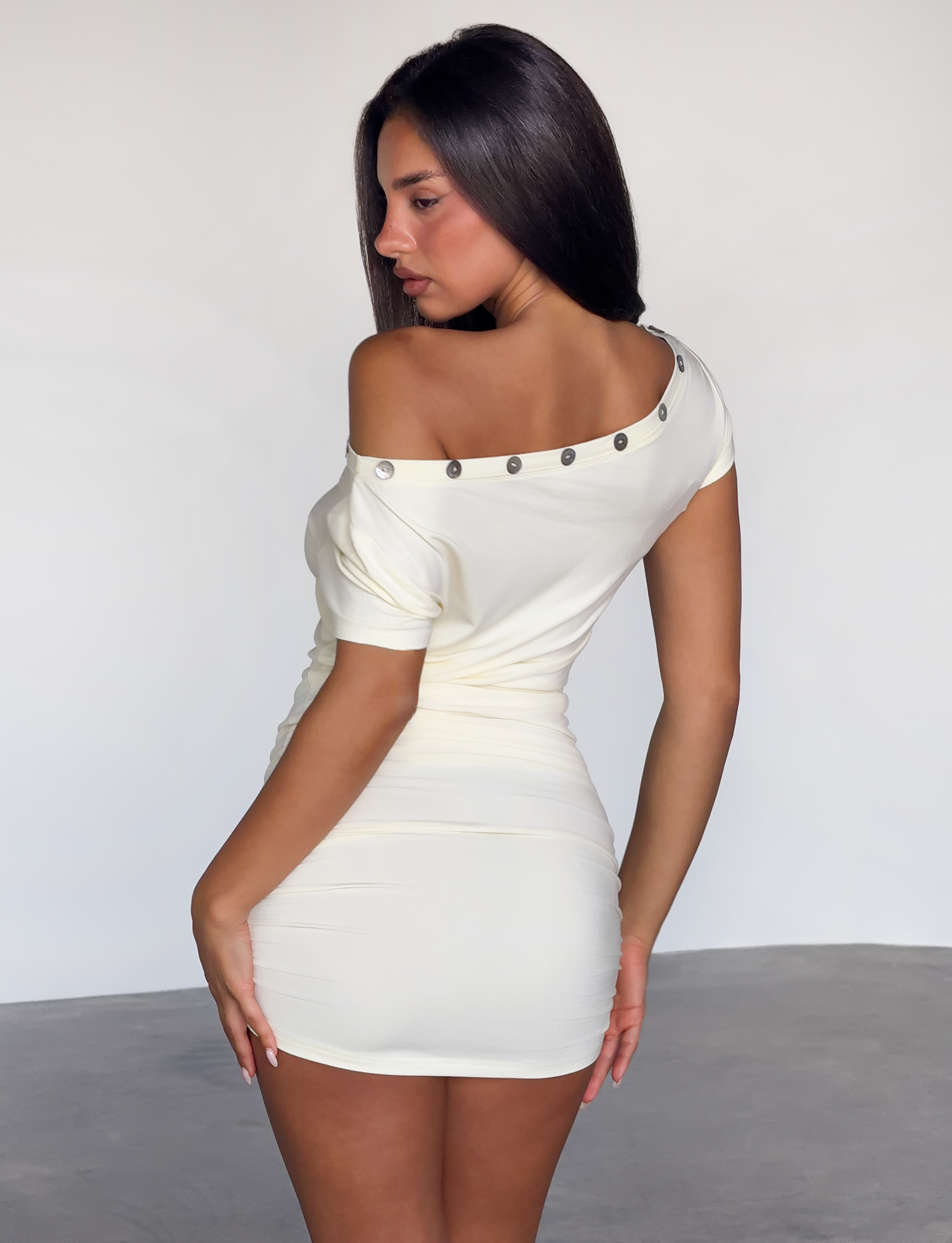 A woman with long dark hair poses in a minimalistic room, wearing the TIGERMIST MARITA MINI DRESS in cream—a fitted off-shoulder style featuring metallic buttons along the neckline, turned to show her back and profile.