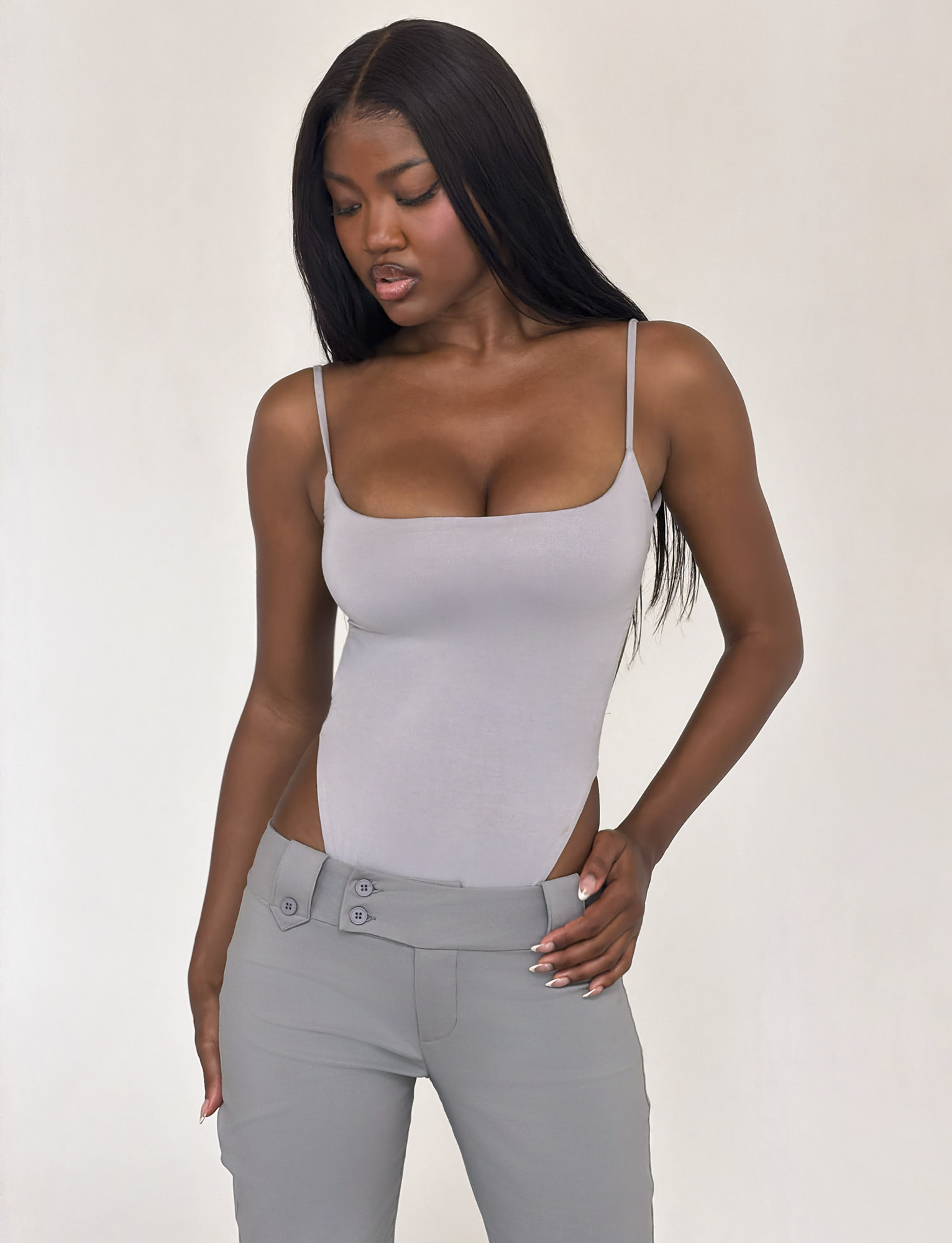 A woman with long straight hair models the TIGERMIST MINERVA BODYSUIT in frost grey with high-waisted grey pants. She looks down and to the side, hand on her hip, posing against a plain background.