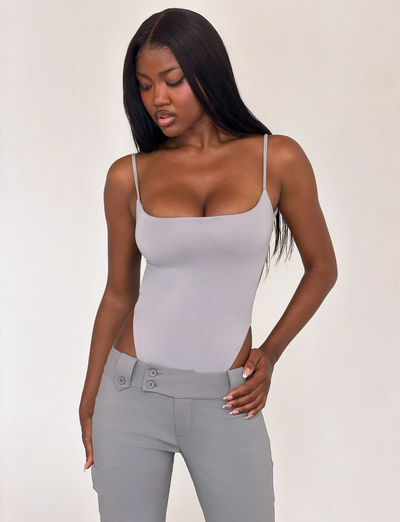 A woman with long straight hair models the TIGERMIST MINERVA BODYSUIT in frost grey with high-waisted grey pants. She looks down and to the side, hand on her hip, posing against a plain background.