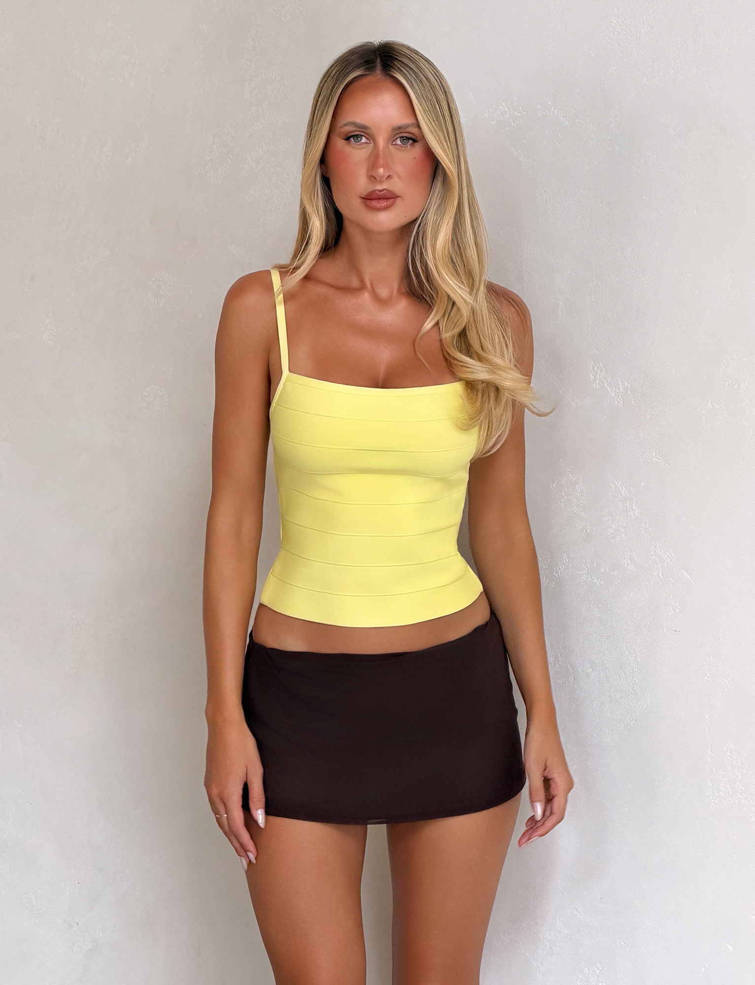 A woman with long blonde hair wears the TIGERMIST MIDORI BANDAGE TOP in yellow and a short brown skirt, standing against a light textured wall and looking directly at the camera with a neutral expression.