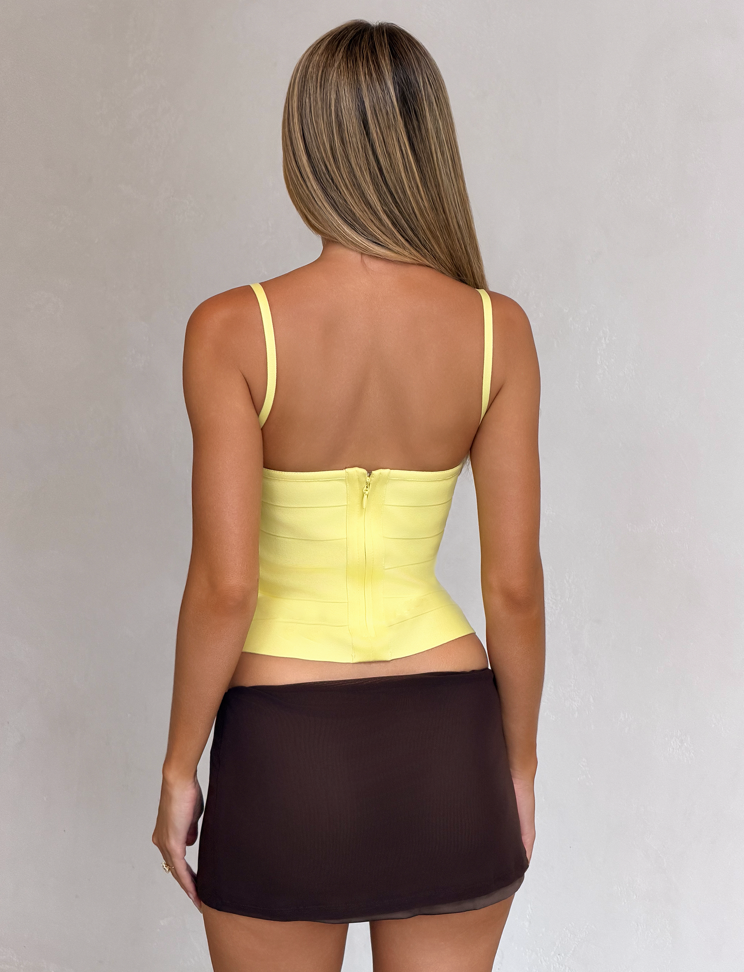 A woman with long, straight hair models the TIGERMIST MIDORI BANDAGE TOP in yellow, featuring a square neckline and thin straps, paired with a black mini skirt. She stands facing away against a plain, light-colored background.