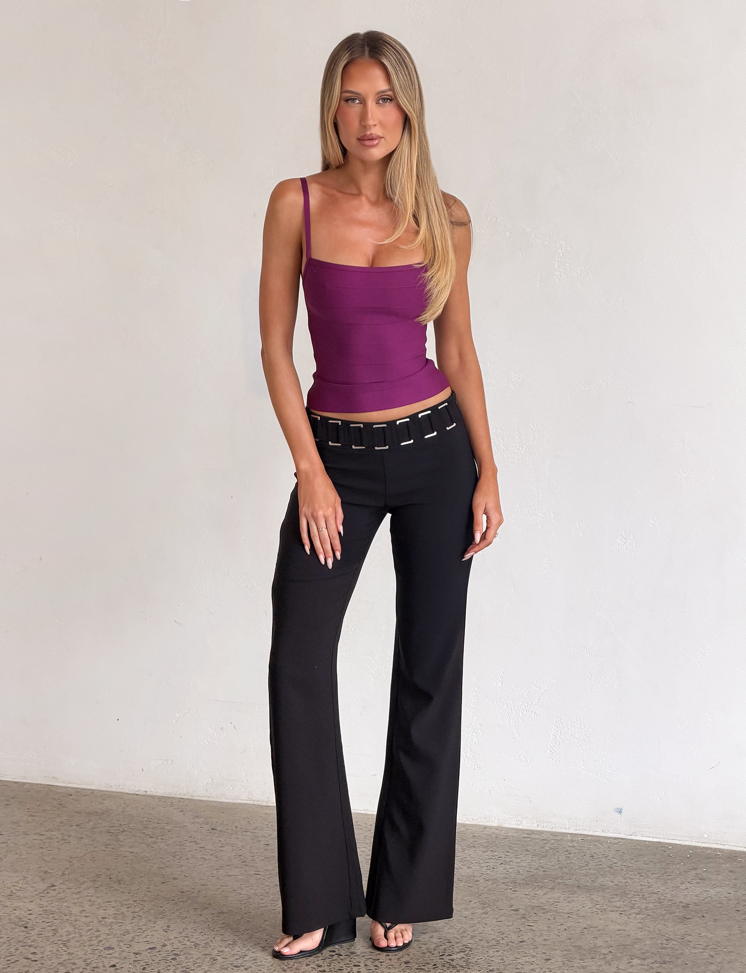 A woman with long blonde hair wears the TIGERMIST MIDORI BANDAGE TOP in plum, featuring a square neckline, paired with belted black pants. She stands indoors against a plain white wall on a concrete floor.