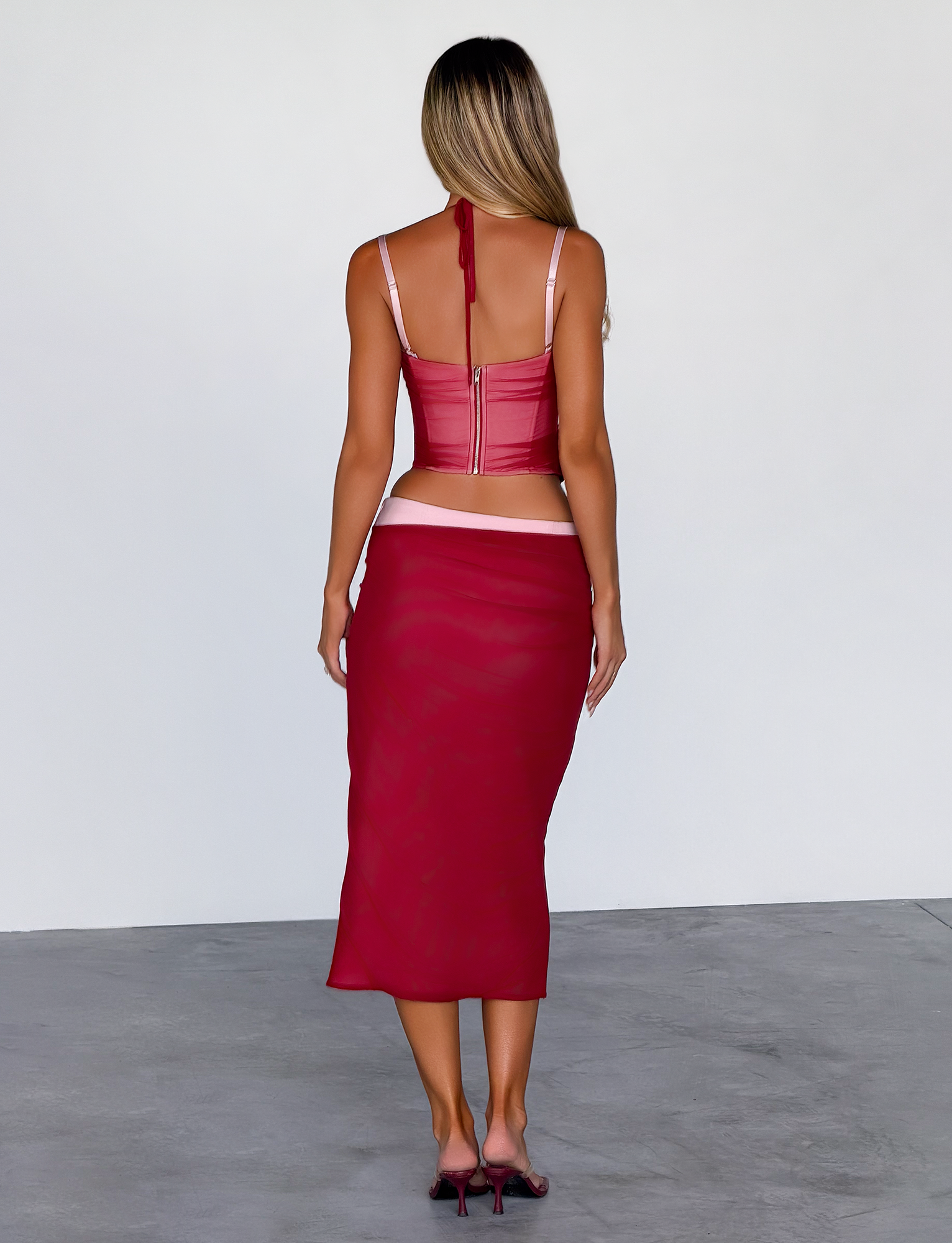 A woman with long, light brown hair stands facing away, wearing the TIGERMIST CALINE MIDI SKIRT in red and a matching crop top with thin straps, paired with red heels against a plain, light background.