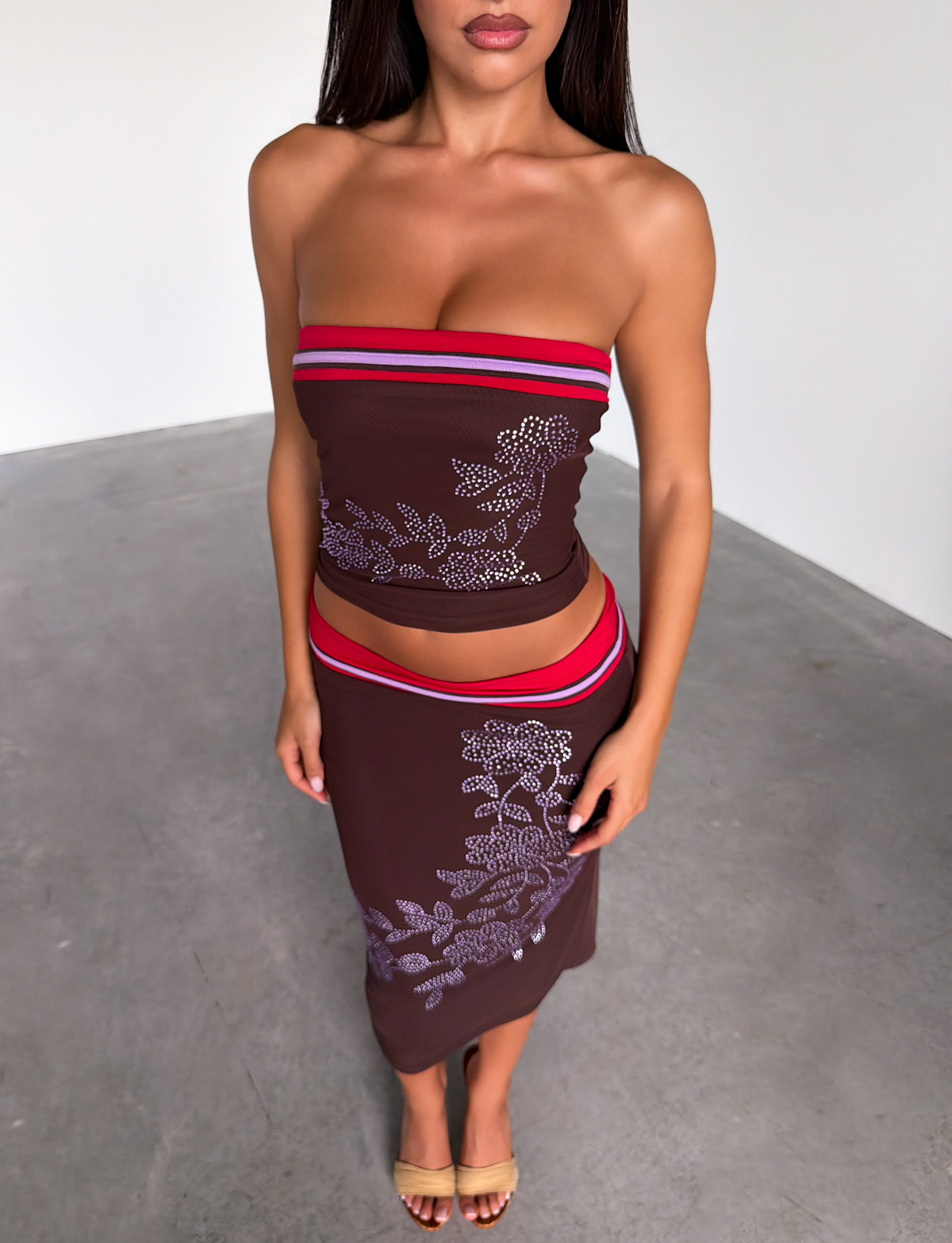A woman stands indoors wearing the TIGERMIST SERAH DIAMANTE BOOB TUBE in chocolate with a matching skirt featuring red stripes and white floral patterns, paired with tan heeled sandals. The background is plain with gray flooring and white walls.