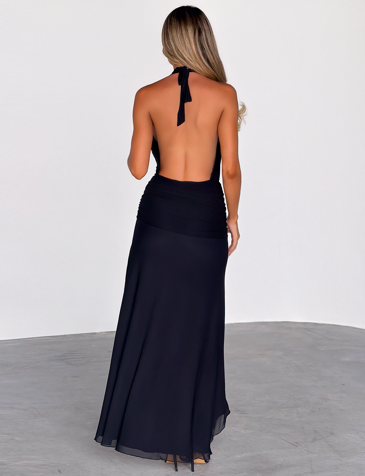 A woman with long, wavy hair stands on a gray floor facing away, wearing the TIGERMIST CORELLE DRESS - BLACK, a backless halter neck maxi dress that flows to the ground and contrasts elegantly with the white background.