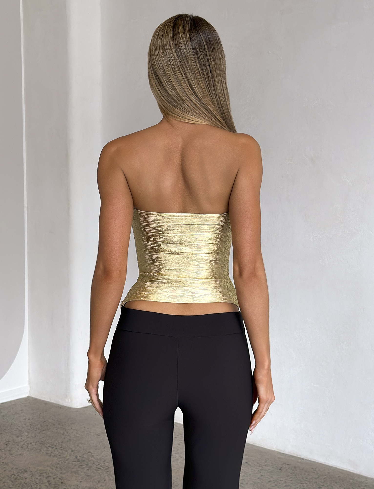 A woman with straight, light brown hair stands facing away in a minimalist room with light-colored walls and a speckled floor, wearing the TIGERMIST NIYA METALLIC BANDAGE TOP in gold paired with black pants.