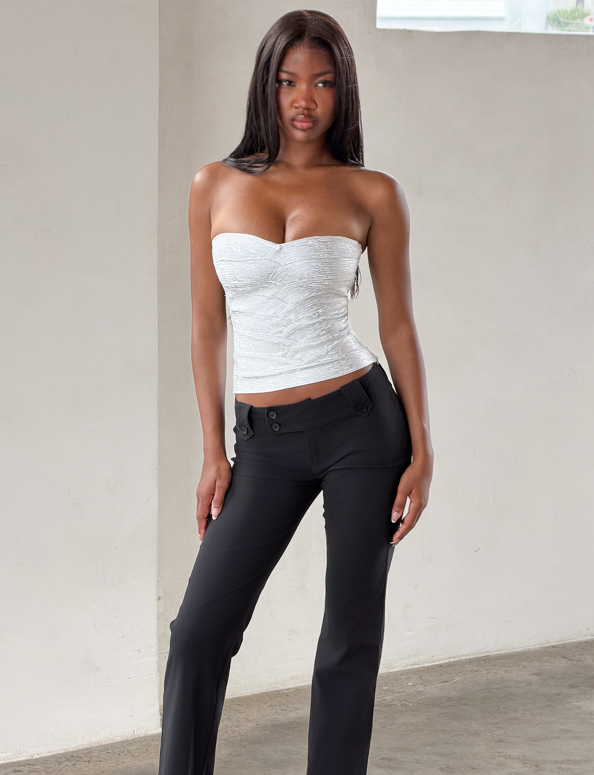A woman stands indoors against a light wall, wearing the TIGERMIST NIYA METALLIC BANDAGE TOP in Liquid Silver and black pants. She has long straight hair and gazes forward with a neutral expression.