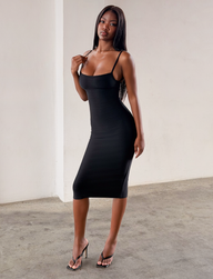A woman wearing the TIGERMIST MIDORI BANDAGE MIDI DRESS in black—a fitted, sleeveless, bodycon midi dress—pairs it with black heels as she stands indoors on a concrete floor, posing with one hand on her shoulder and looking confidently at the camera.