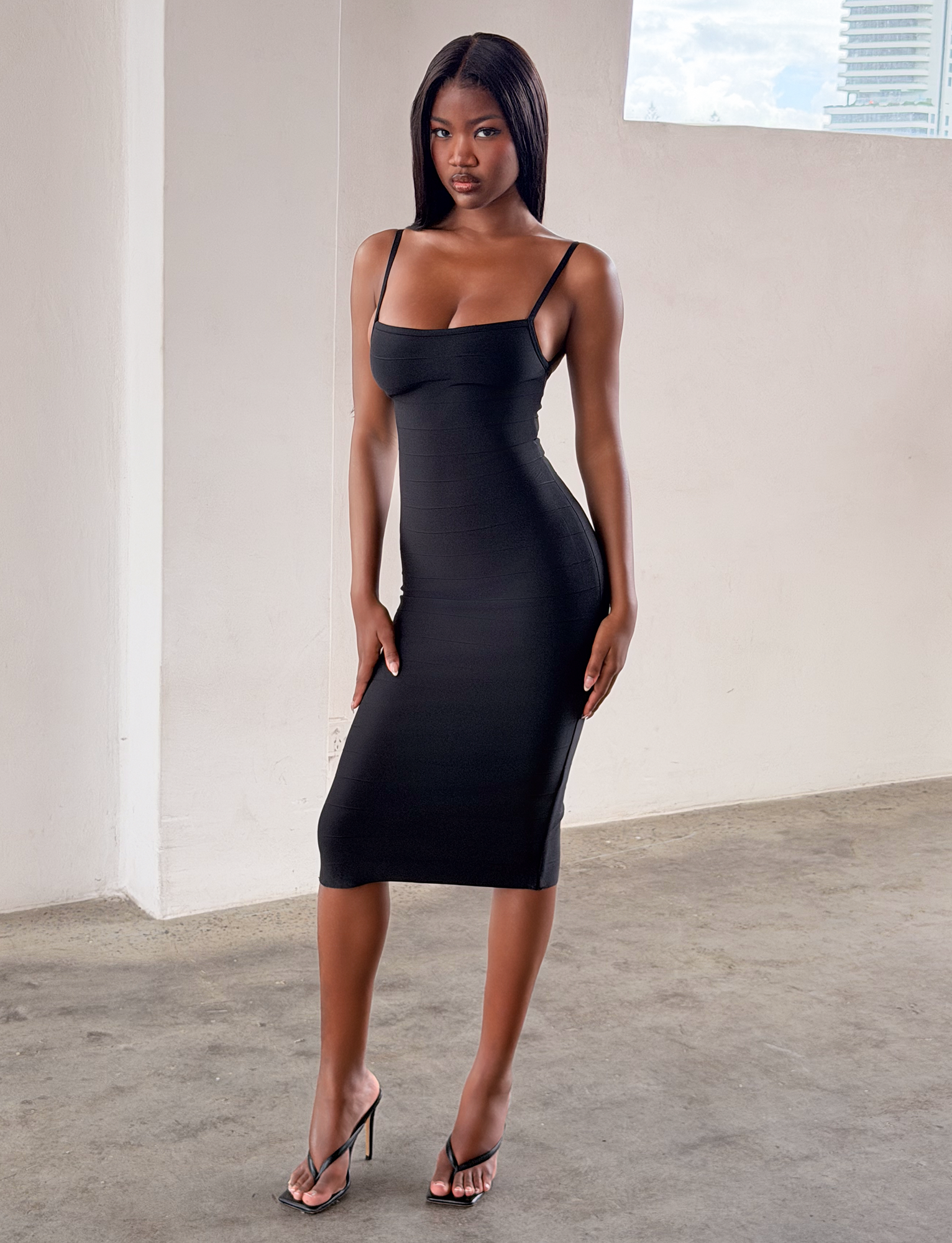 A woman stands indoors on a concrete floor, wearing the TIGERMIST MIDORI MIDI BANDAGE DRESS in black with black heels. Her straight hair frames her neutral expression. A window behind her reveals part of a building and sky.