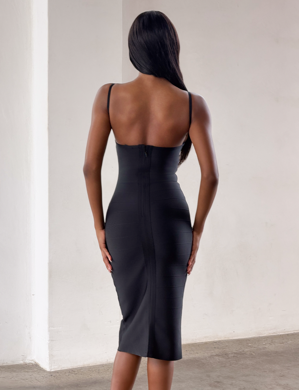 A woman with long dark hair stands indoors, facing away, wearing the TIGERMIST MIDORI BANDAGE MIDI DRESS in black—a fitted, sleeveless dress with thin straps that hugs her body in a classic bodycon fit. The room features plain white walls and a concrete floor.