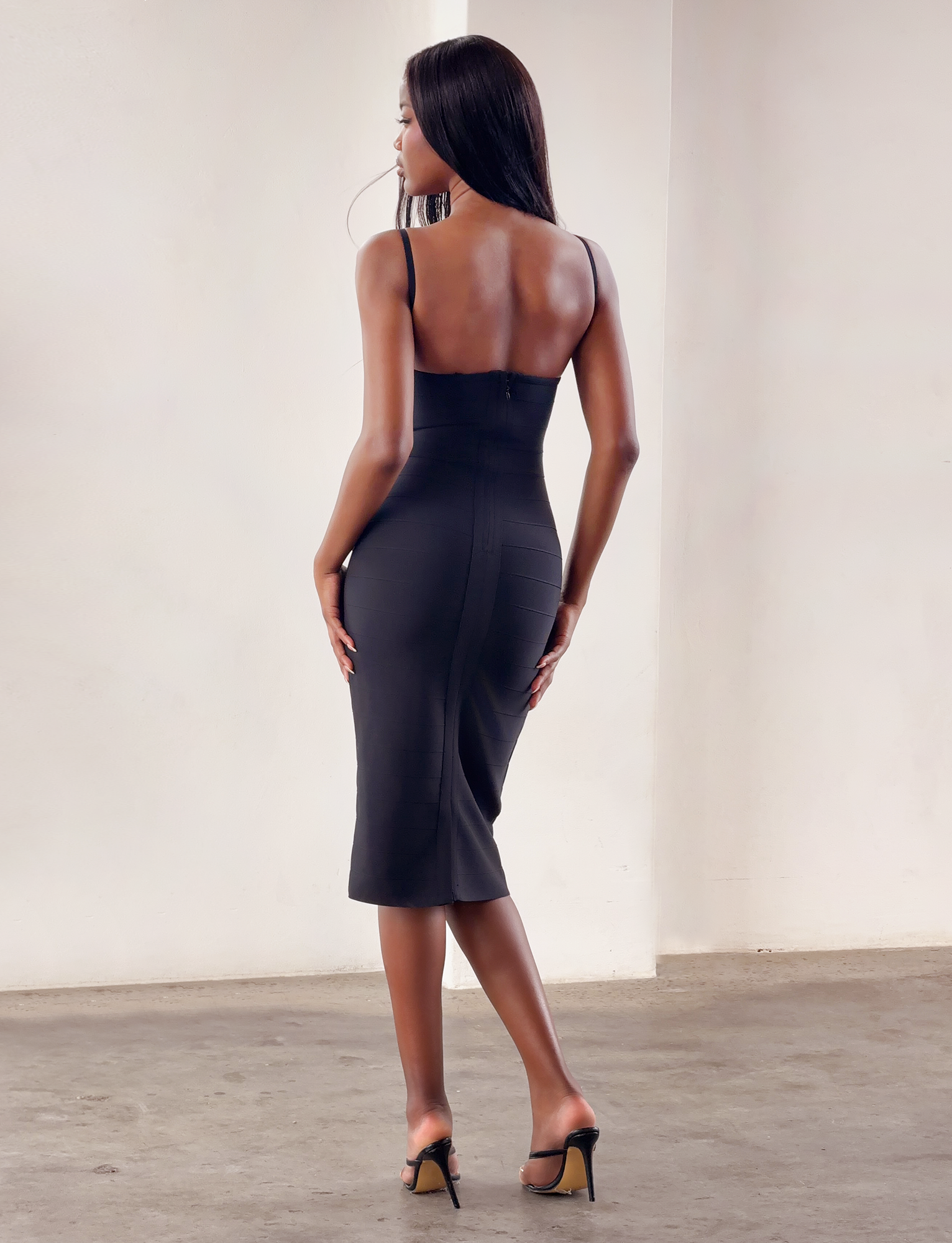 A woman with long straight hair stands with her back to the camera in a minimalist room, wearing the TIGERMIST MIDORI MIDI BANDAGE DRESS in black and black high-heeled shoes.
