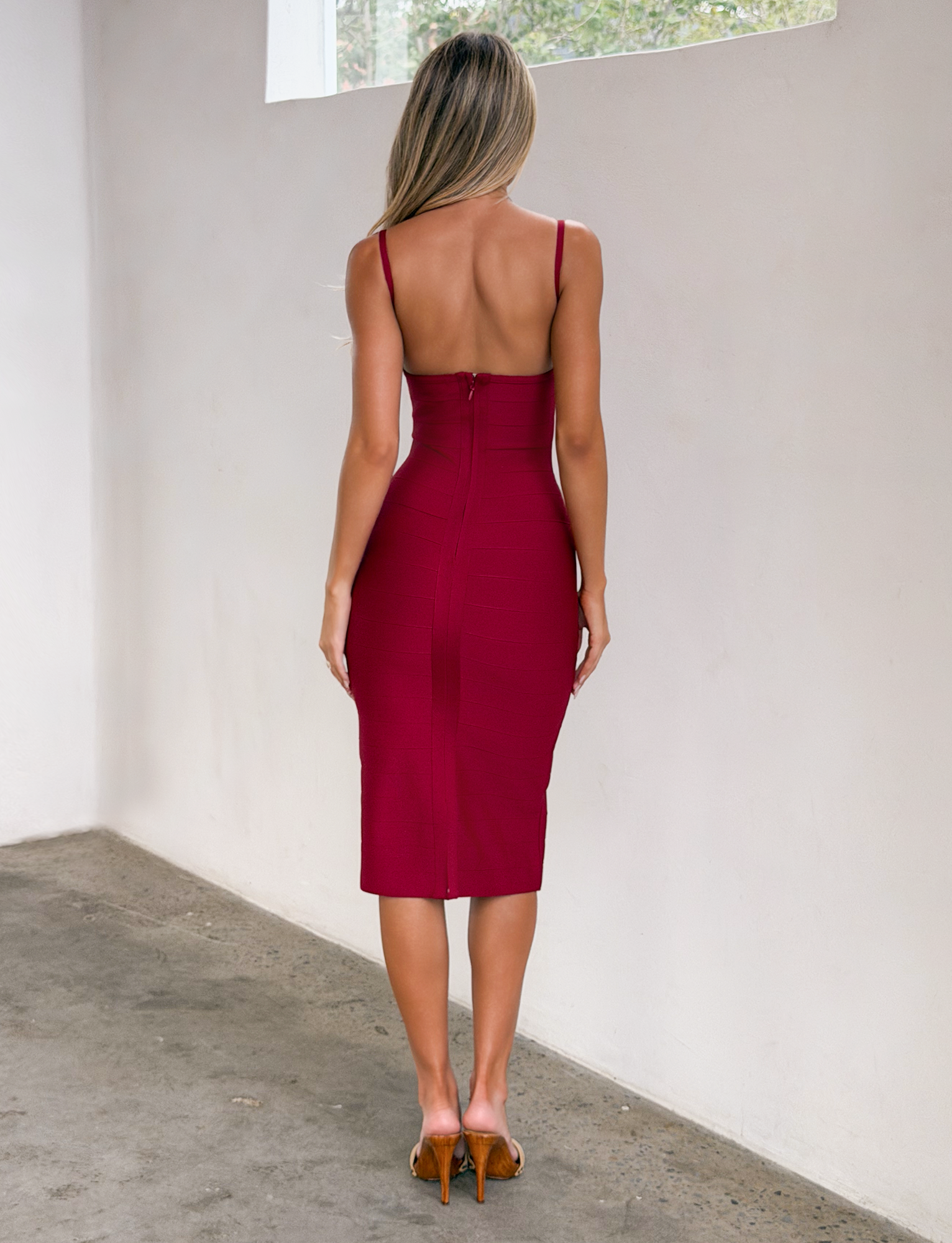 A woman with long blonde hair stands indoors, facing away, wearing the TIGERMIST MIDORI MIDI BANDAGE DRESS in cherry red and tan heels. The room features white walls, a window, and a concrete floor.