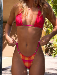 A woman outdoors adjusts her bikini bottoms while wearing the TIGERMIST AYANNA REVERSIBLE BIKINI TOP in Gingham and Resort Plaid. Her face is not visible.
